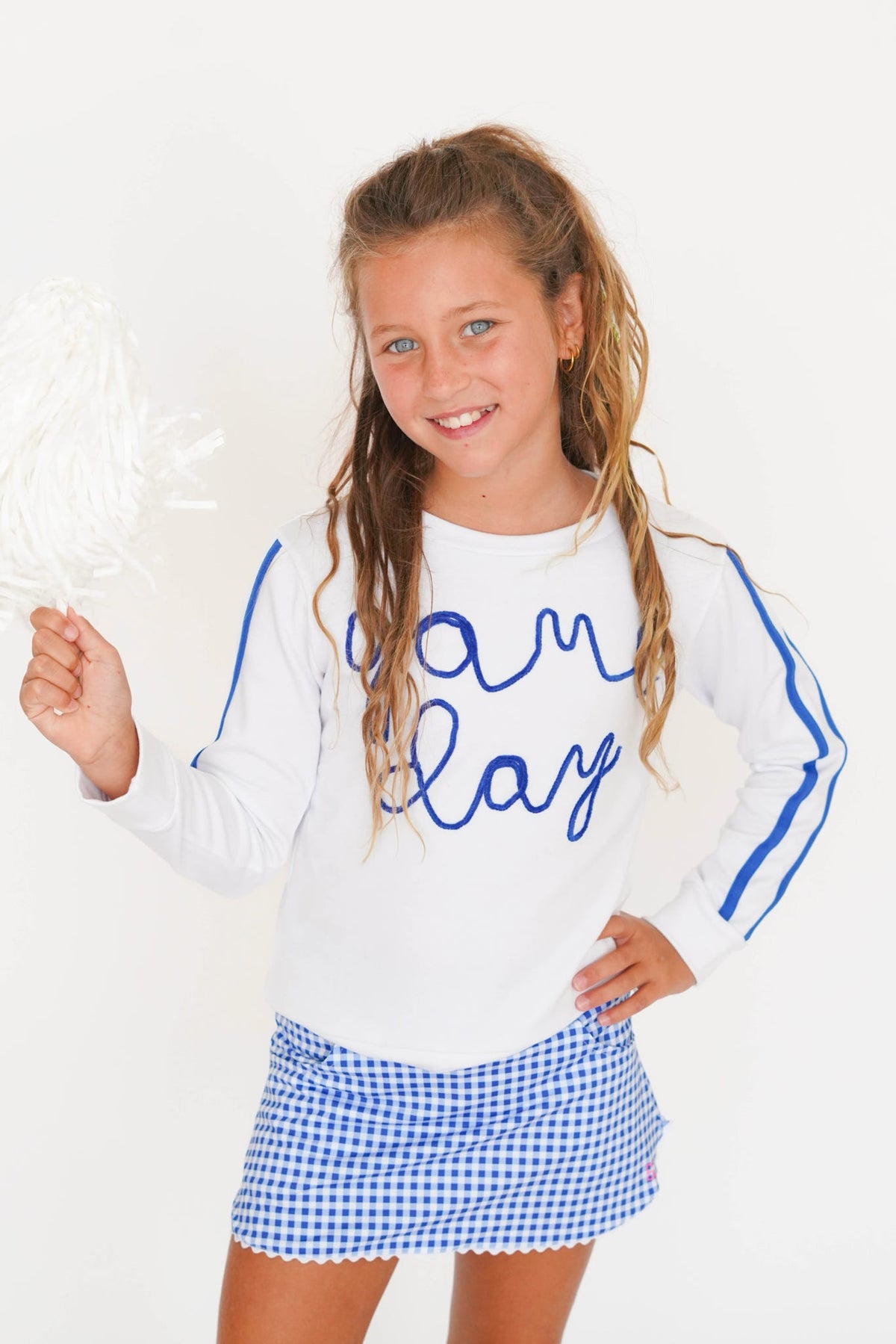 A young girl with blonde hair in a ponytail smiles while holding a white pom-pom. She is wearing a white long-sleeve shirt with blue script reading 'I am Clay' and blue stripes on the sleeves, paired with a blue and white gingham mini skirt, against a plain white background.