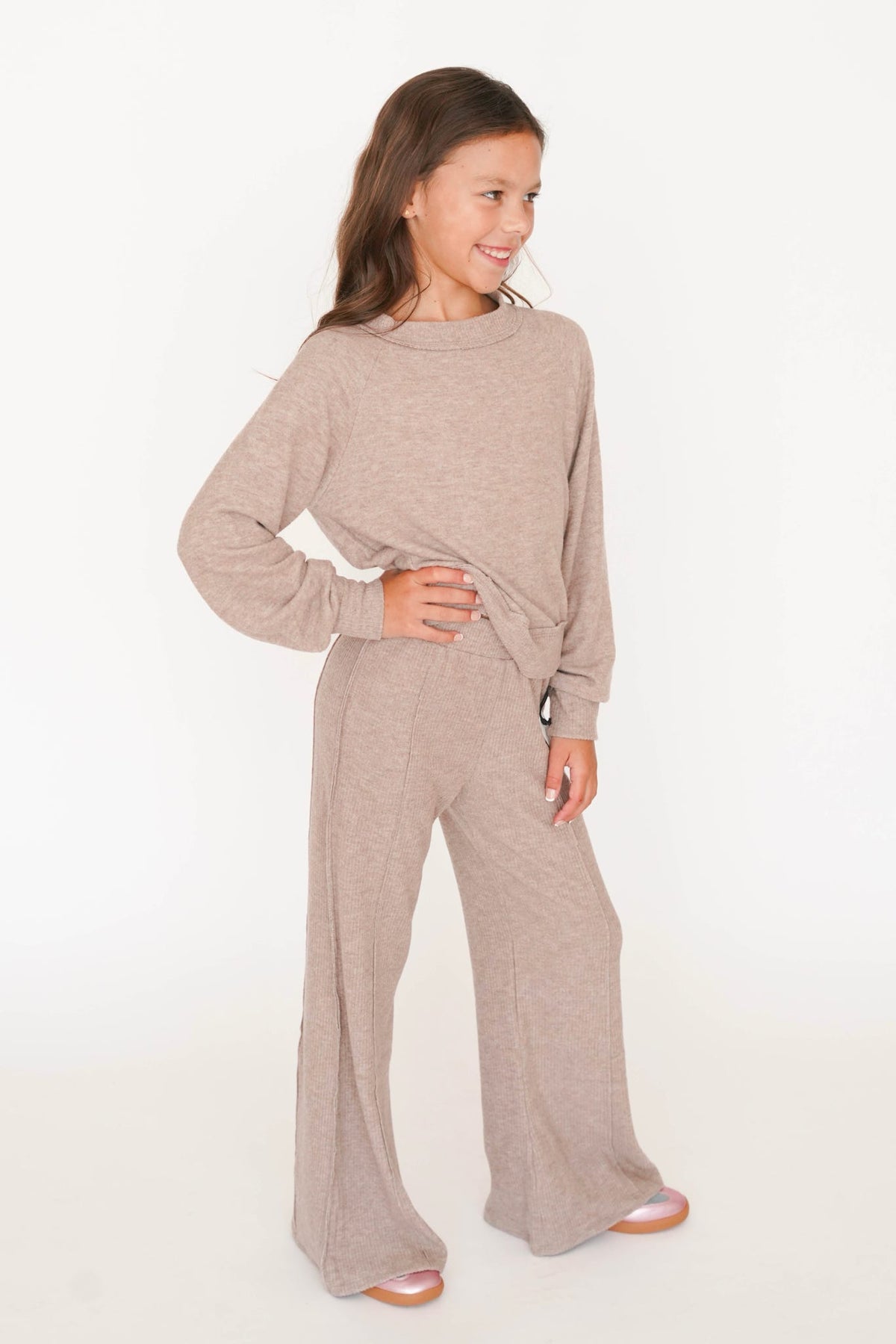 A young girl modeling a matching two-piece loungewear set consisting of a long-sleeve knit top and wide-leg pants in a heathered taupe color, set against a plain white studio background.