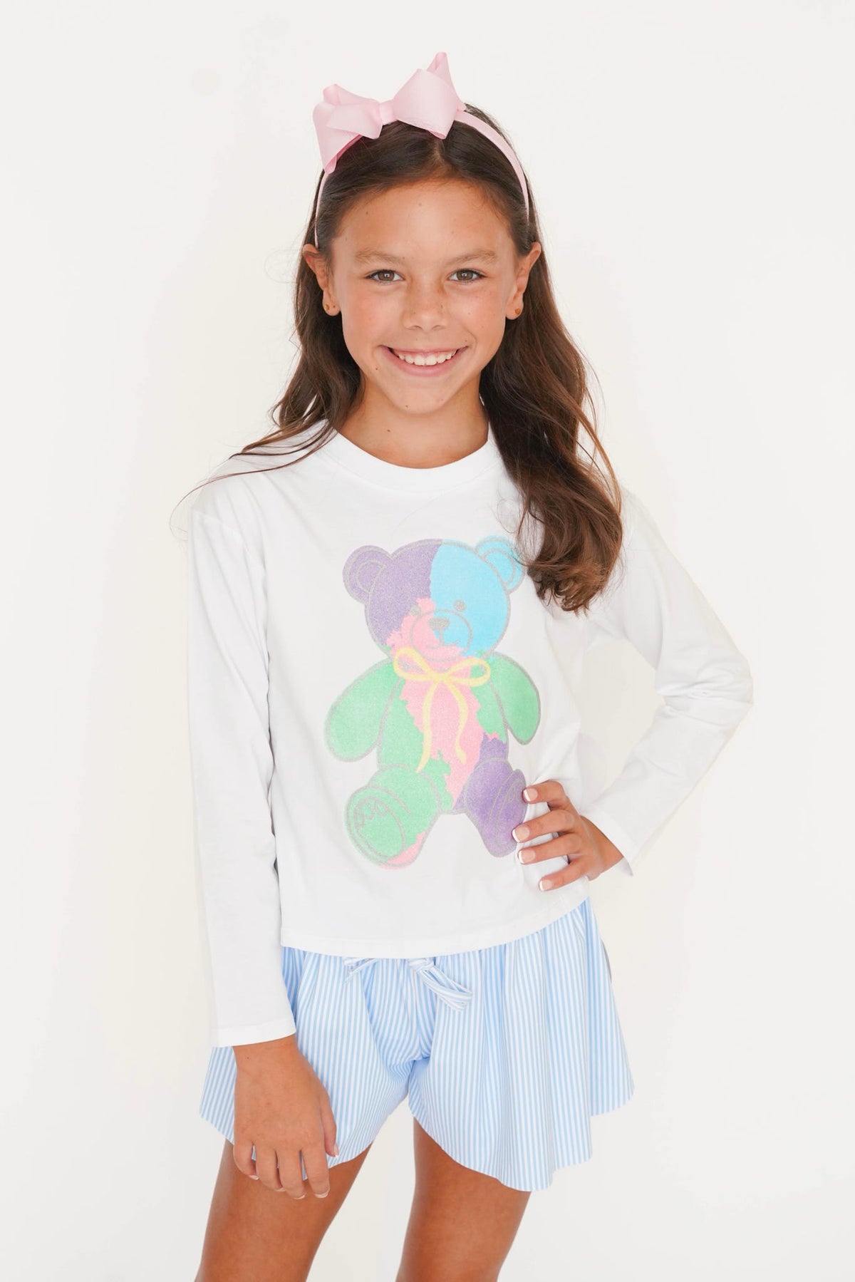 A young girl smiling and posing in a white long-sleeve t-shirt with a colorful pastel teddy bear graphic, paired with light blue and white striped shorts, and a pink bow headband against a plain white background.