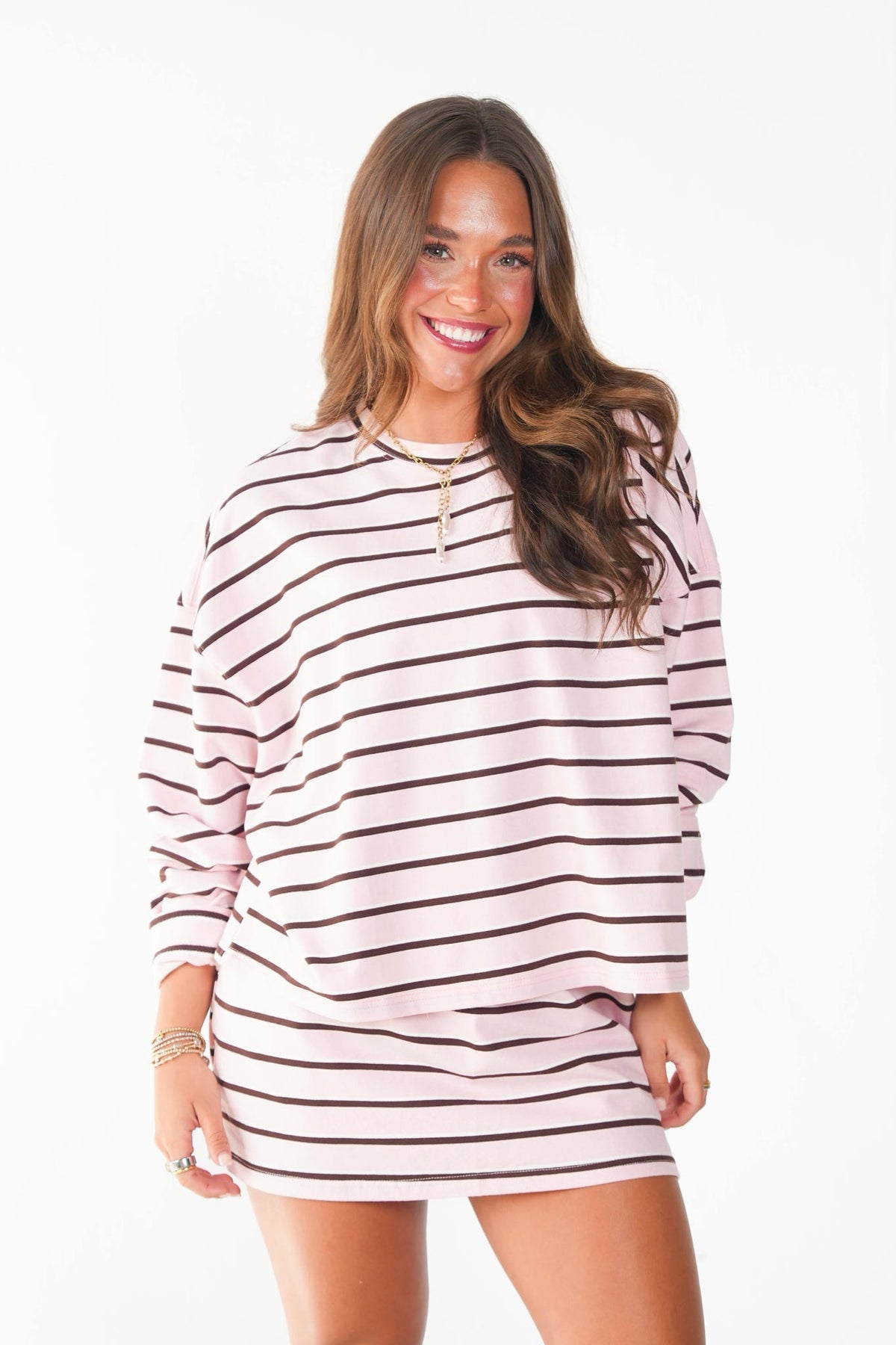 A woman modeling a light pink and dark brown horizontally striped long-sleeve sweatshirt dress against a plain white background.