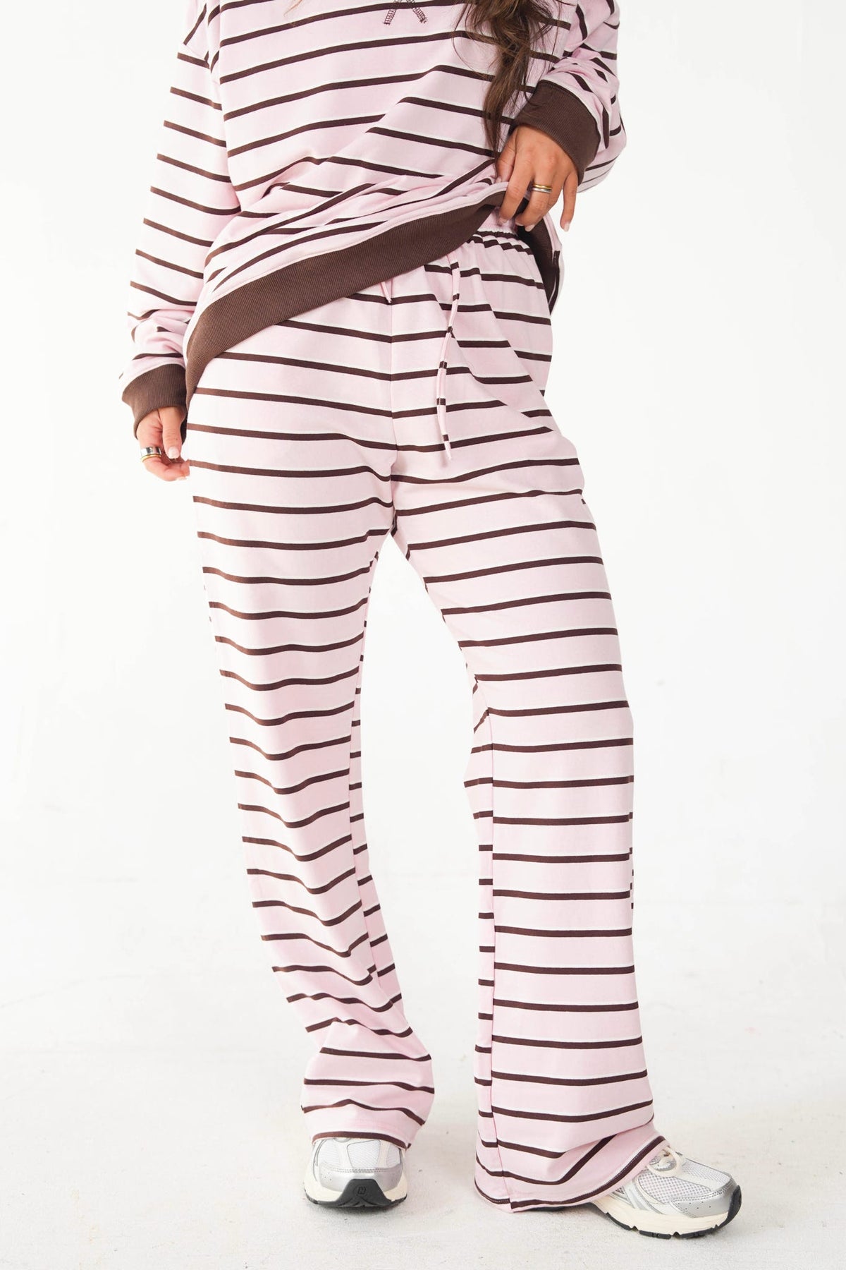 A person wearing a matching lounge set with a light pink and dark brown horizontal stripe pattern, consisting of a long-sleeve top and wide-leg pants, paired with white sneakers against a plain white background.