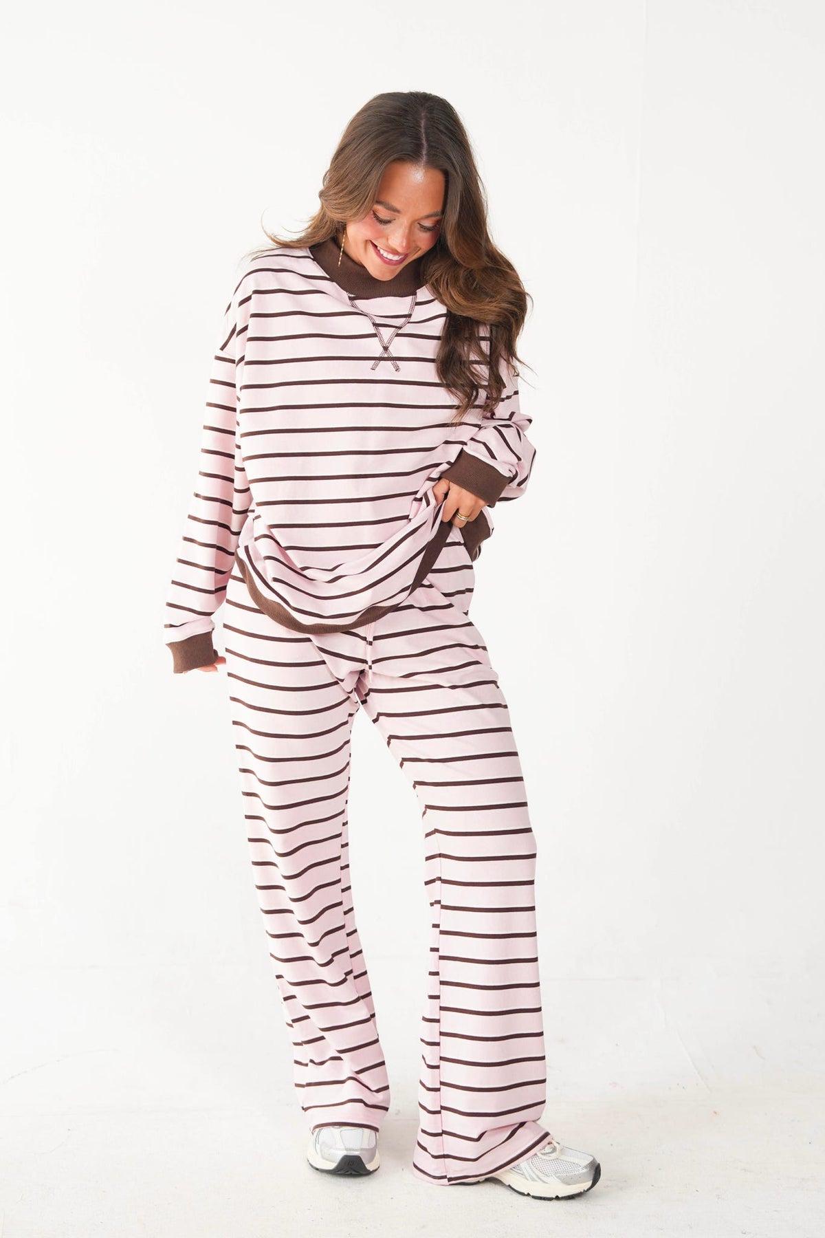 A woman modeling a two-piece long-sleeve pajama set with horizontal stripes in pink and brown, standing against a plain white studio background.