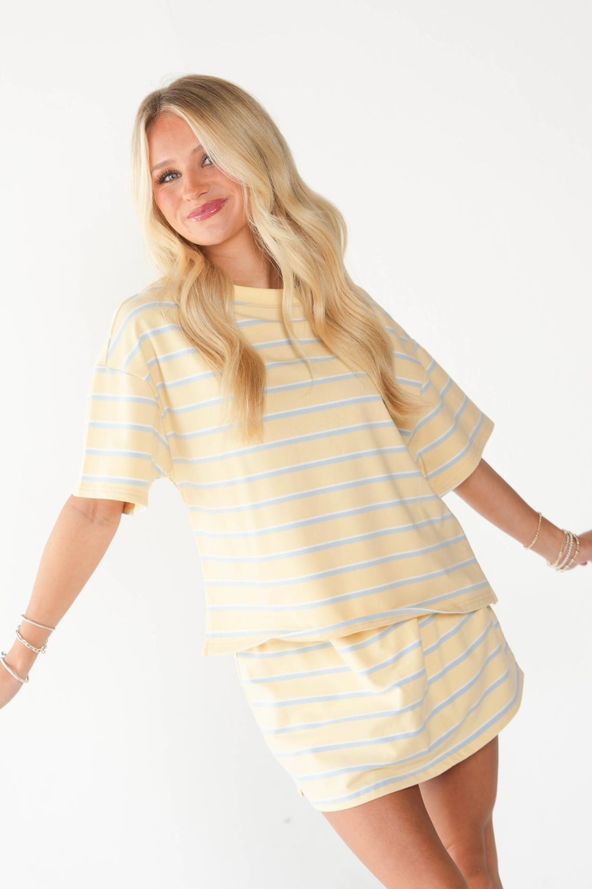 A blonde woman modeling a matching pale yellow and light blue striped lounge set, featuring a short-sleeve top and a short skirt, against a plain white studio background.