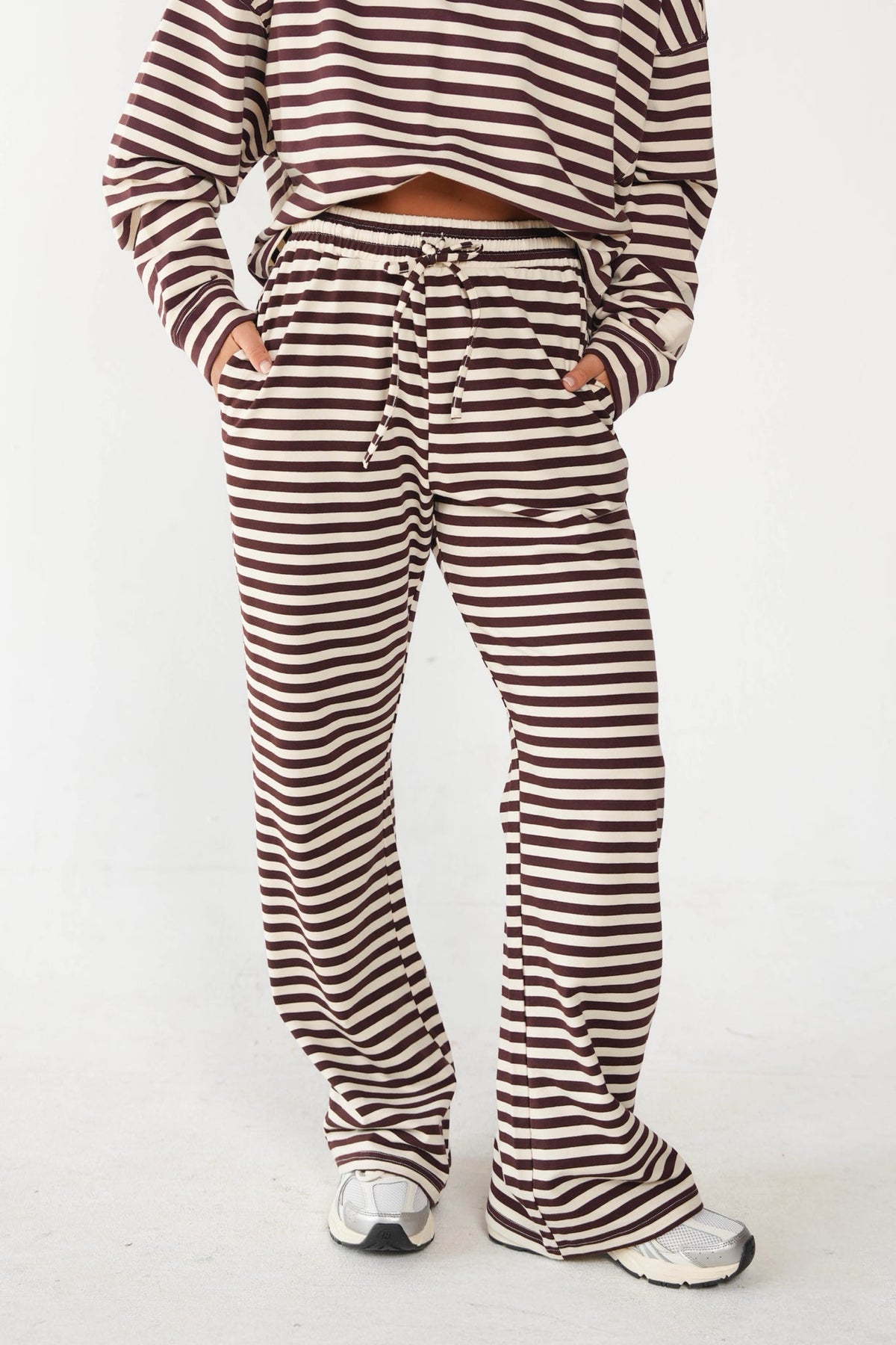 A person wearing a matching brown and cream striped lounge set, featuring a cropped long-sleeve top and wide-leg drawstring pants, paired with white sneakers, against a plain white studio background.