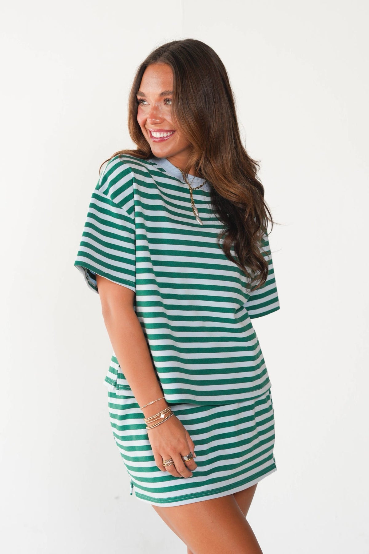 A woman modeling a green and white horizontally striped short-sleeve hooded t-shirt dress against a plain white background.