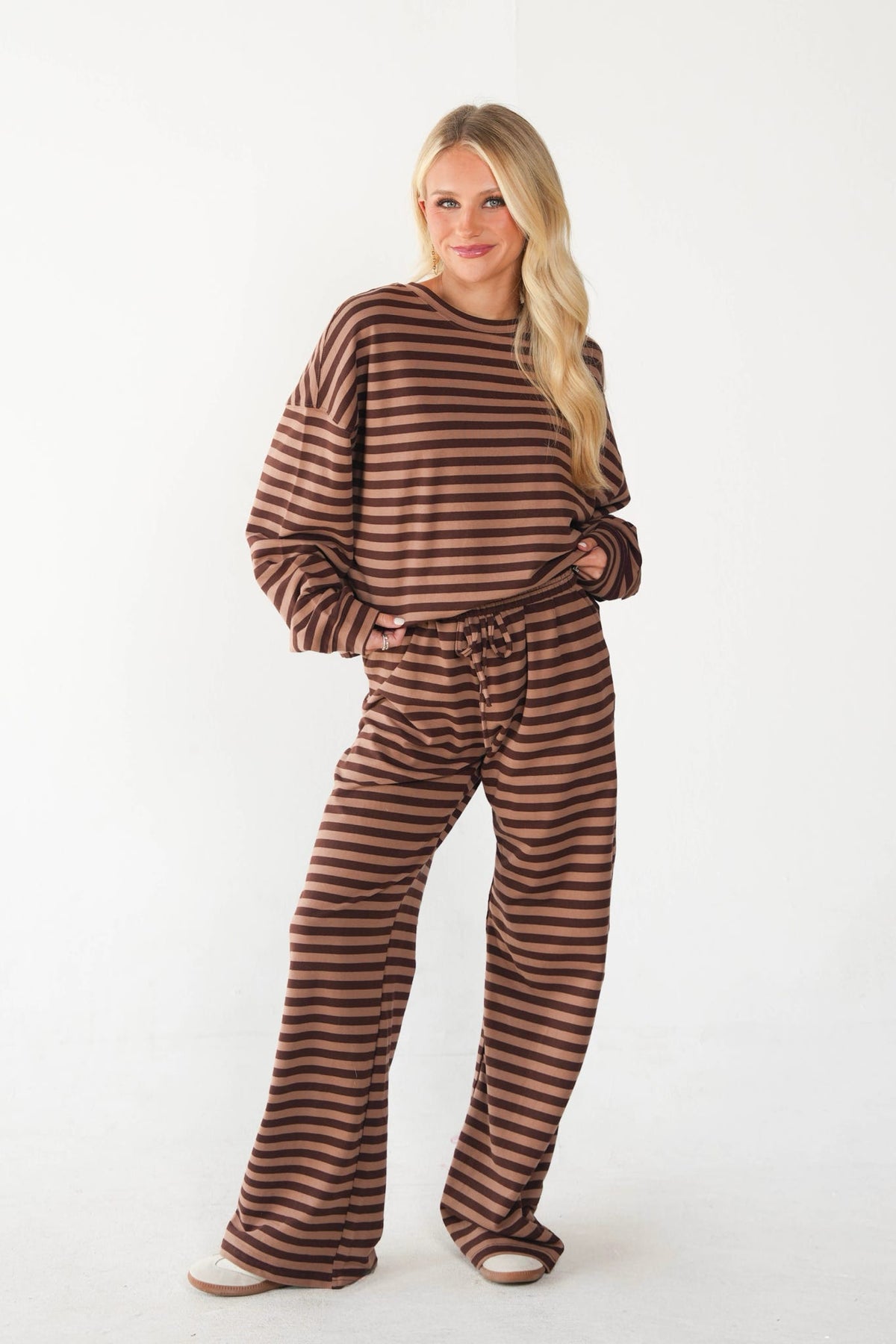 A woman modeling a matching two-piece loungewear set with brown and beige horizontal stripes, consisting of a long-sleeve top and wide-leg pants, against a plain white studio background.