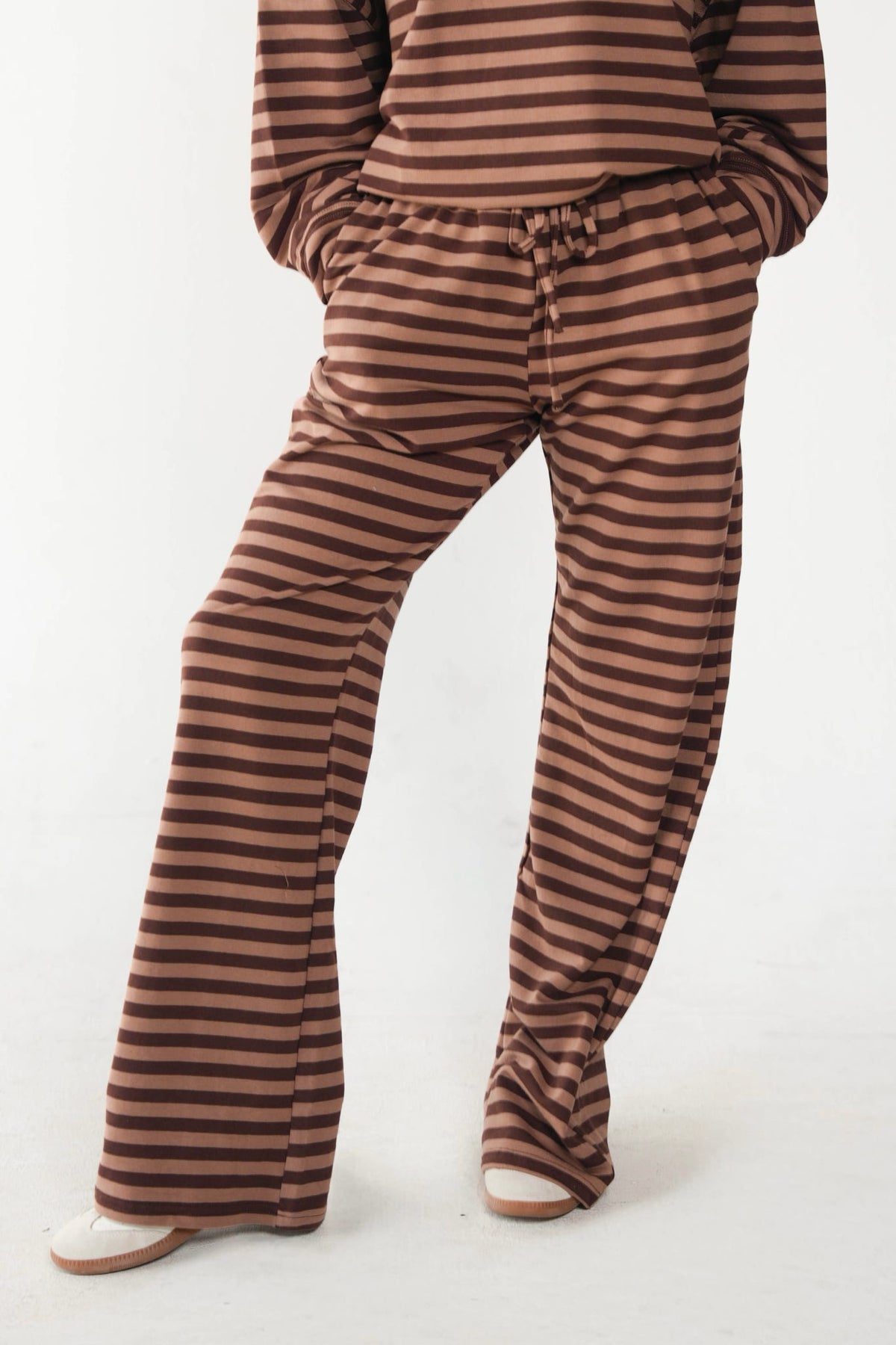 A person wearing brown and tan horizontally striped lounge pants with a drawstring waist, paired with a matching striped top and white slip-on shoes, shown against a plain white studio background.
