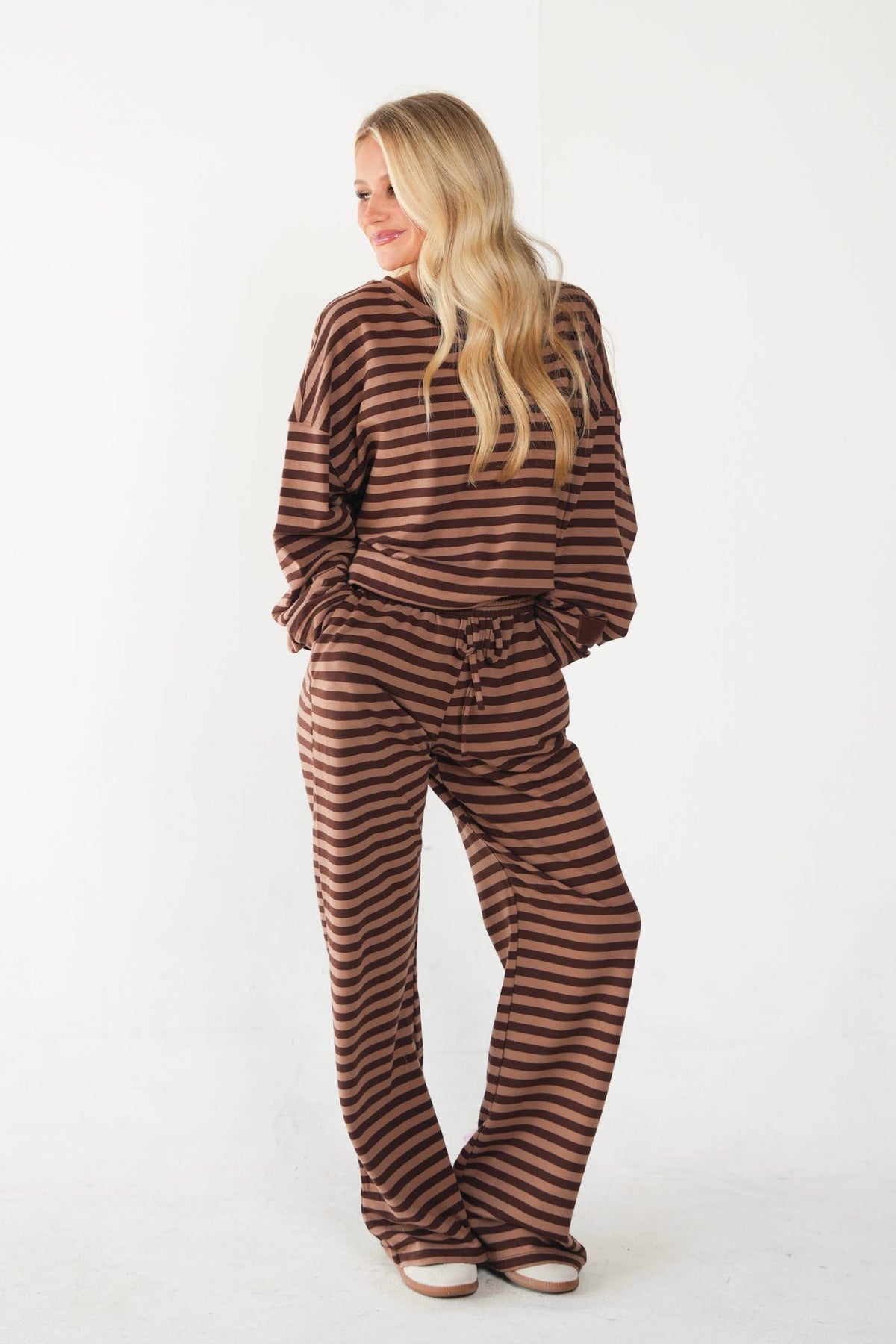 A woman modeling a matching brown and dark brown horizontally striped lounge set, consisting of a long-sleeve top and wide-leg pants, against a plain white studio background.