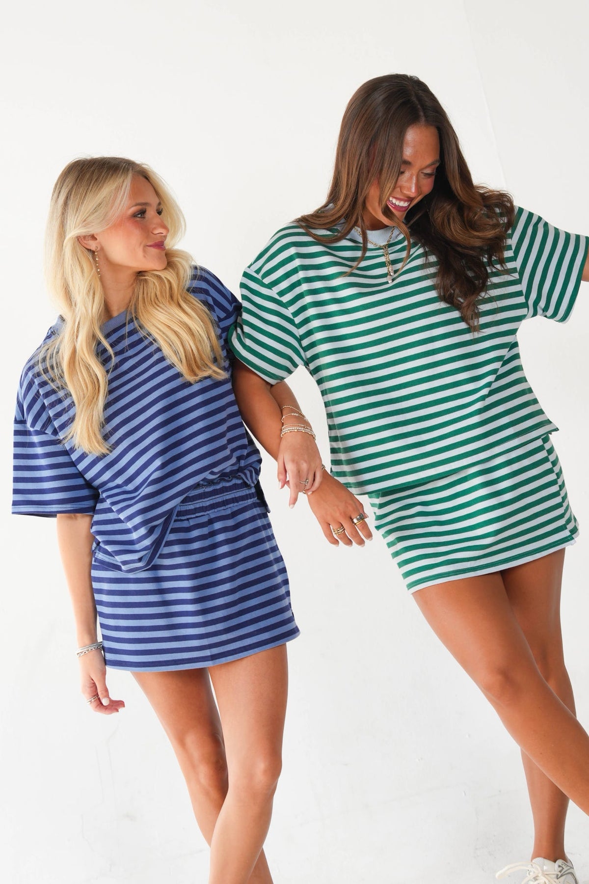 Two women modeling oversized striped t-shirt dresses in a studio setting. The woman on the left wears a blue and white striped dress, and the woman on the right wears a green and white striped dress.