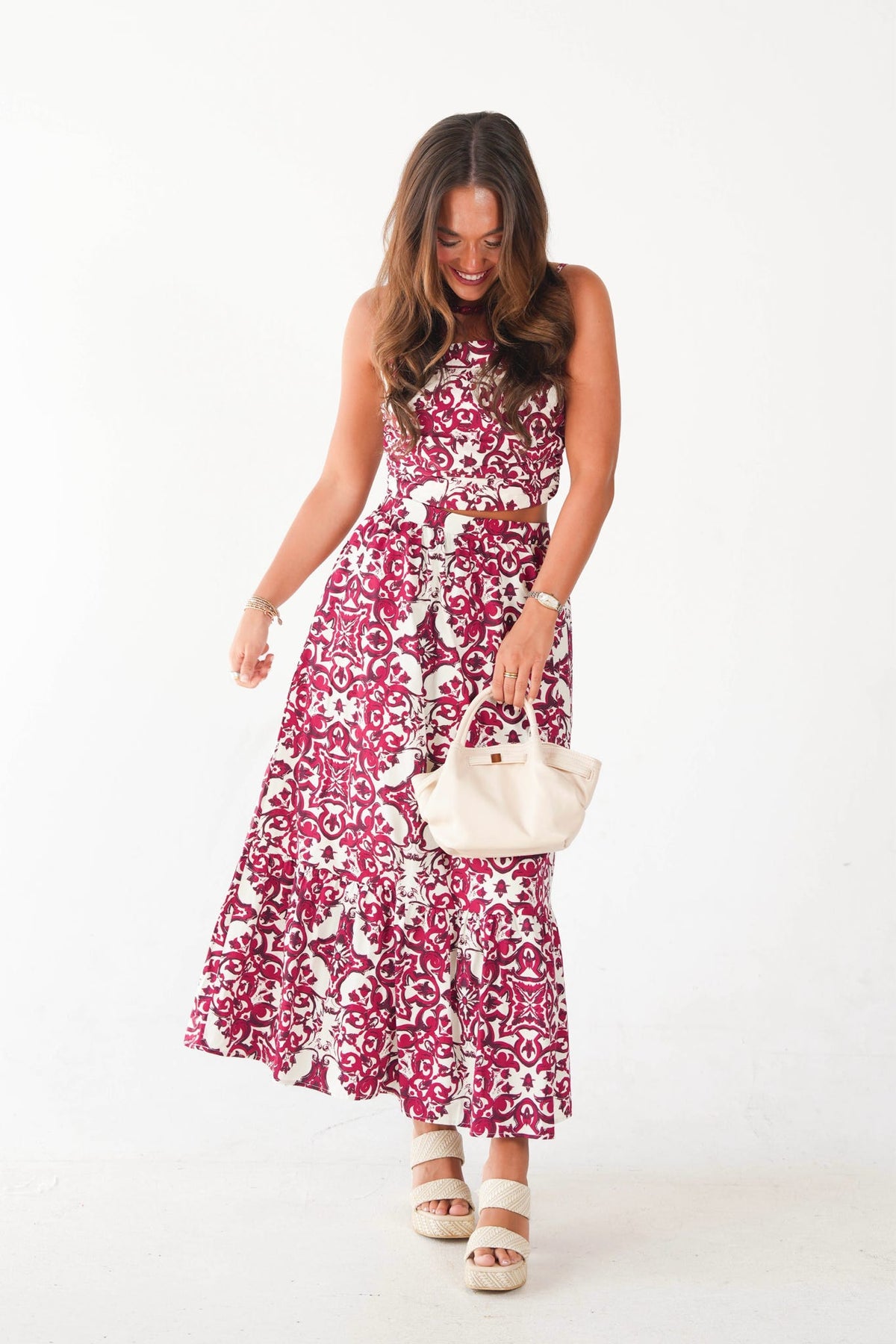 A woman modeling a sleeveless two-piece set featuring a matching halter top and maxi skirt in a magenta and white floral pattern, holding a cream-colored handbag against a plain white studio background.