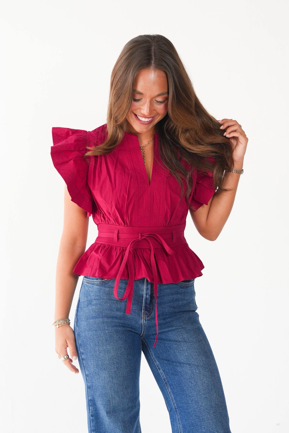 A woman wearing a bright magenta ruffled short-sleeve top with a tie waist and blue jeans, standing against a plain white background.