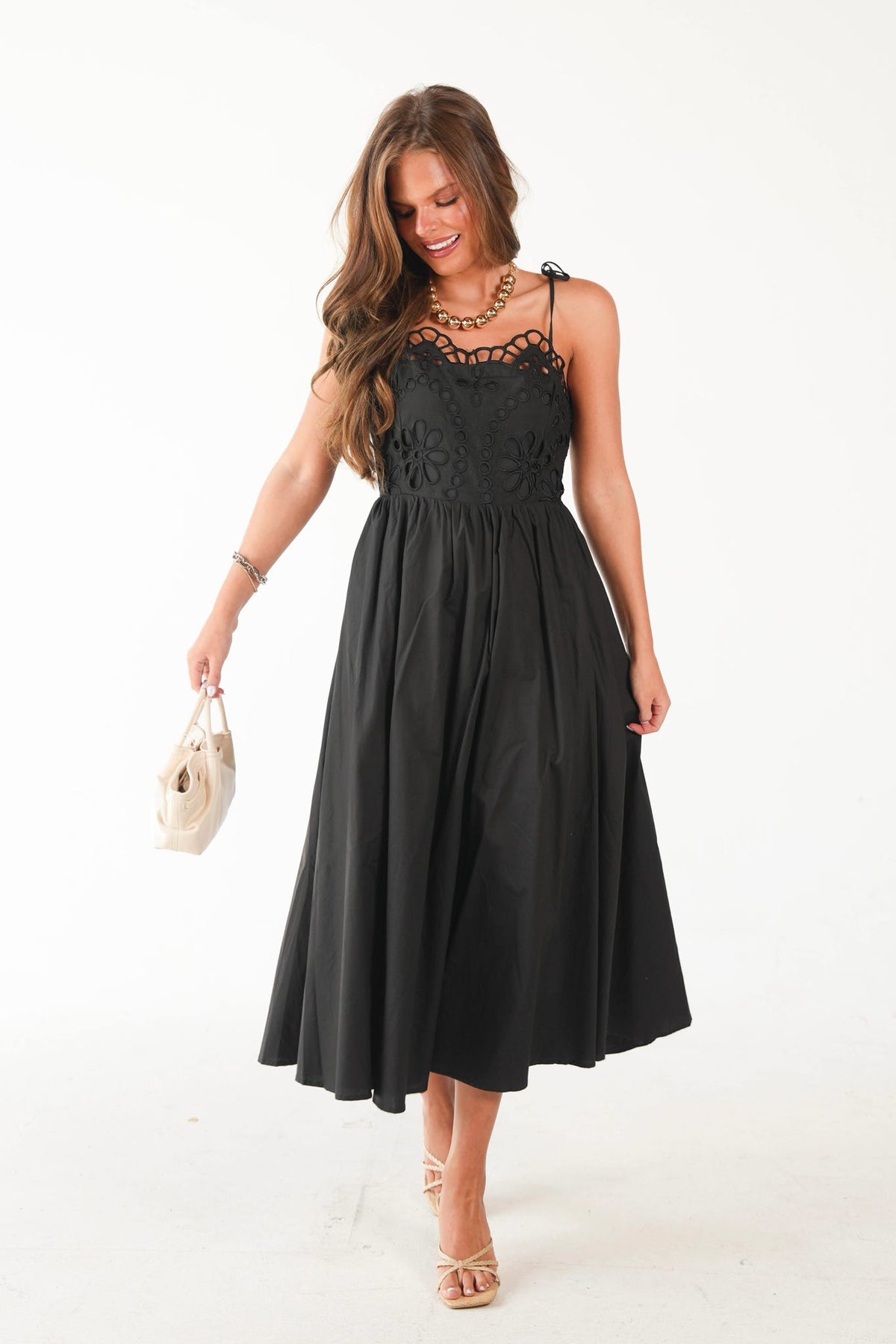A woman modeling a black midi dress with a lace bodice and thin straps, holding a small beige handbag against a plain white studio background.