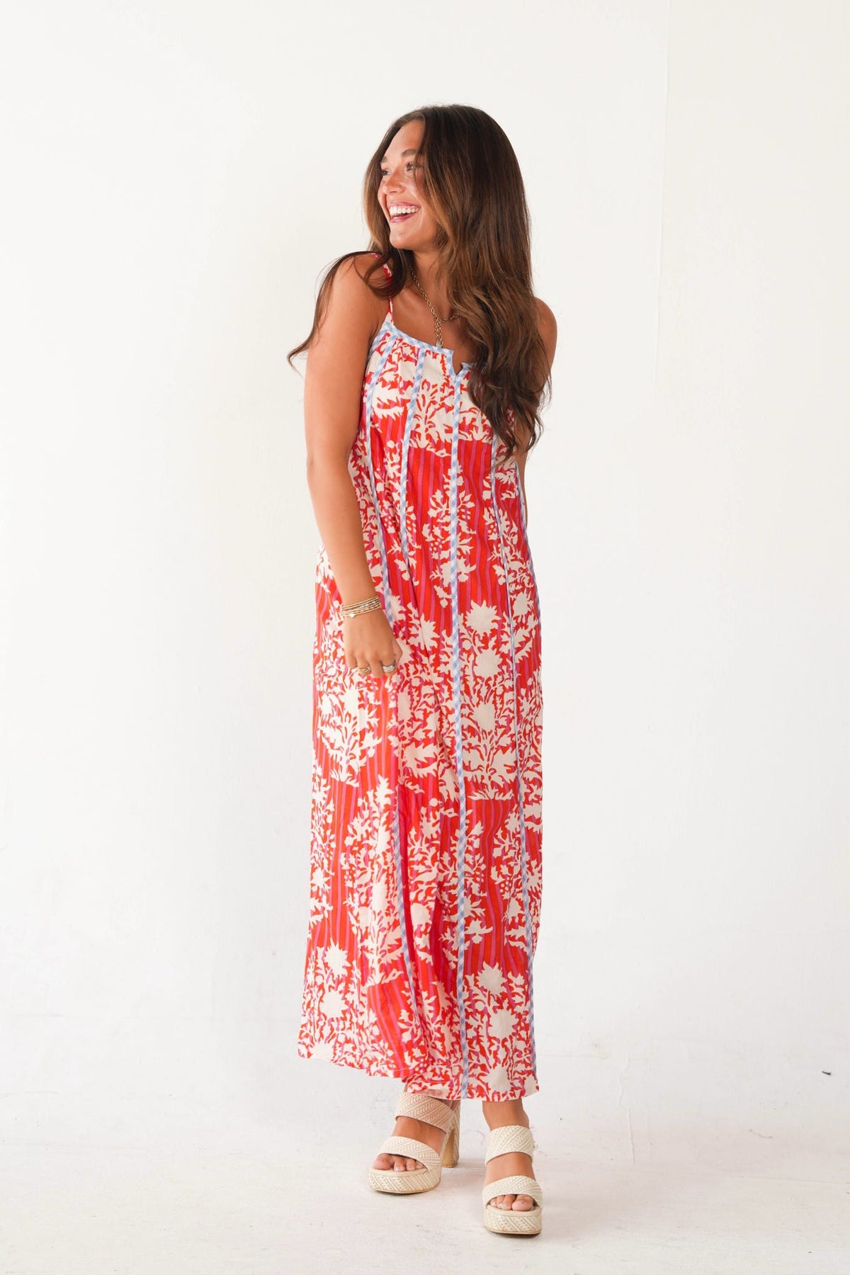 A woman modeling a sleeveless red and white floral maxi dress with a tie-front closure, paired with beige platform sandals, against a plain white studio background.