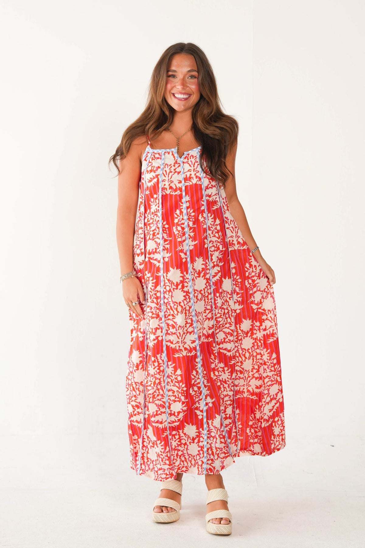 A woman modeling a sleeveless red and white floral maxi dress with a smocked bodice, photographed against a plain white studio background.