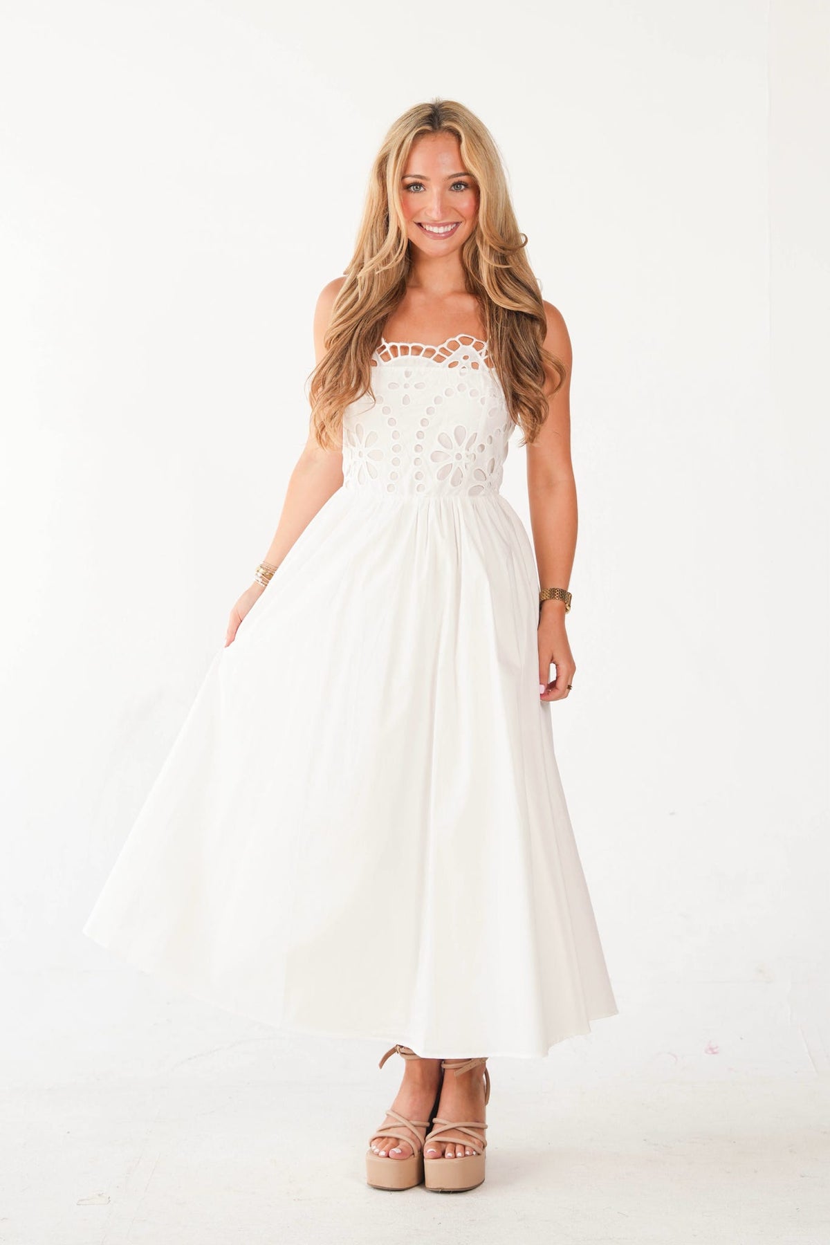 A woman modeling a white strapless maxi dress with a lace bodice and a flowing skirt, paired with beige platform sandals, against a plain white studio background.