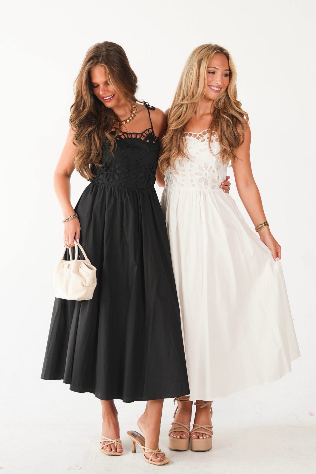 Two women modeling midi-length dresses side by side against a plain white studio background. The woman on the left wears a black sleeveless dress with a lace-trimmed bodice and holds a small cream handbag.