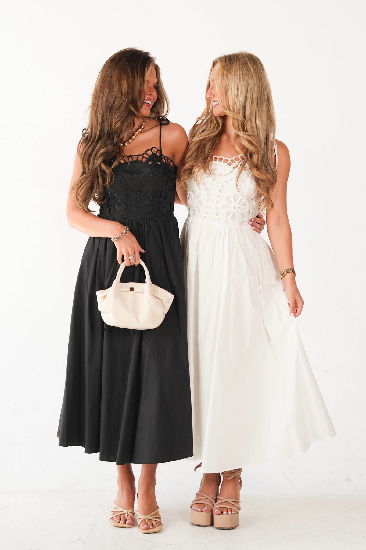 Two women modeling sleeveless midi and maxi dresses against a white studio background. The woman on the left wears a black dress with a lace bodice and holds a cream handbag.