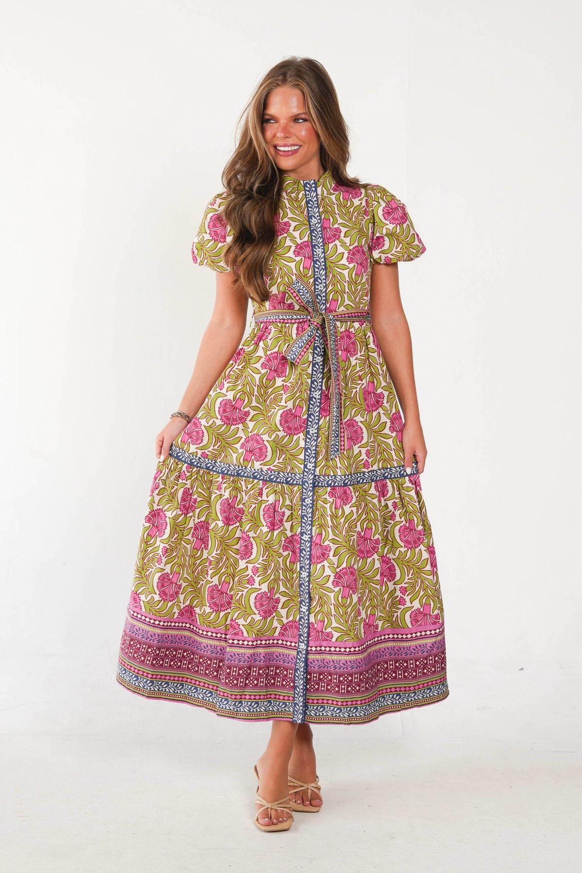 A woman modeling a short-sleeved midi dress with a vibrant floral print in pink, green, and yellow, featuring a matching waist tie and a patterned border hem, set against a plain white studio background.