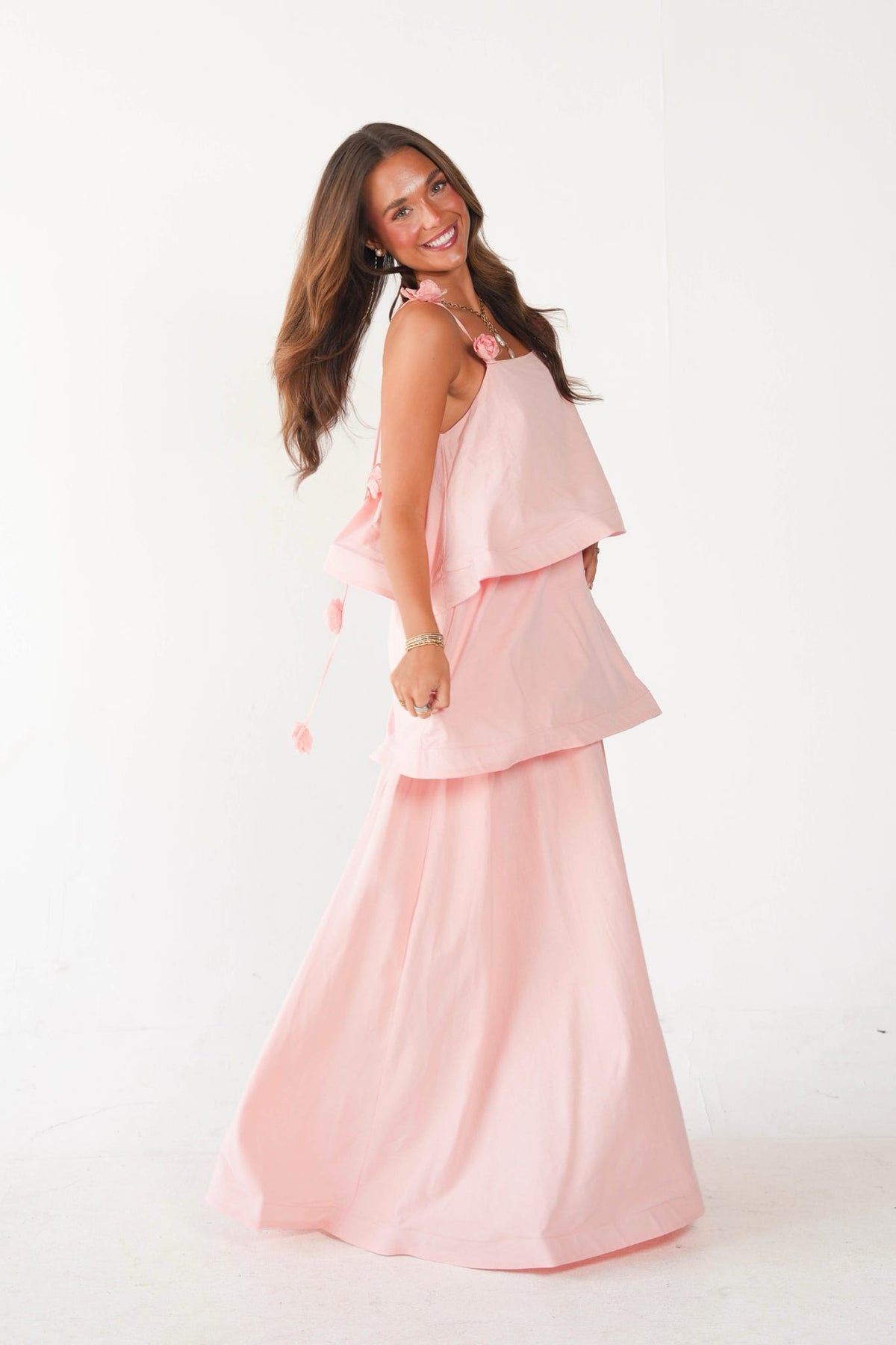 A woman modeling a light pink two-piece outfit consisting of a sleeveless top with layered ruffle sleeves and a matching long skirt, set against a plain white studio background.