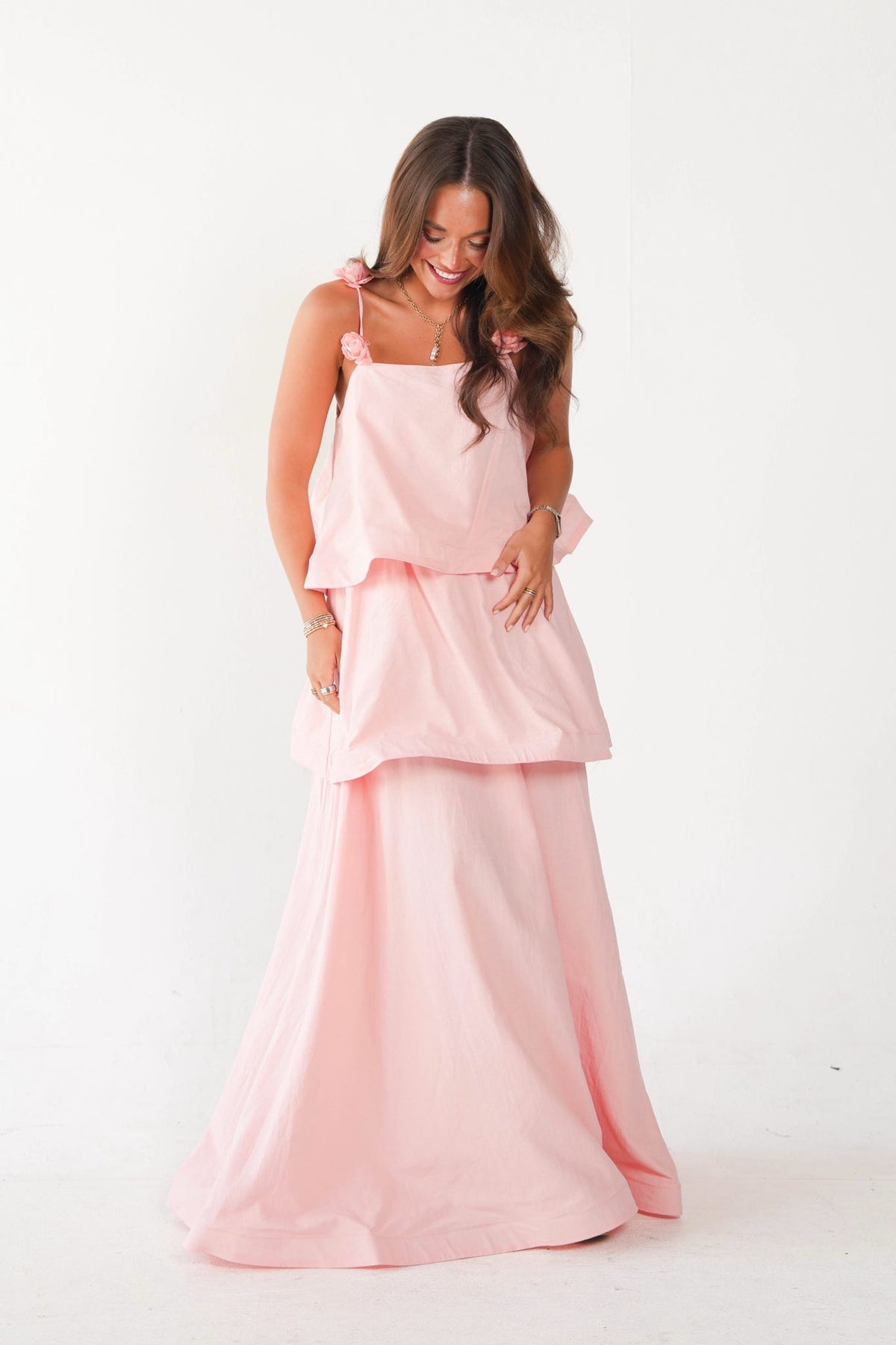 A woman modeling a light pink two-piece outfit consisting of a sleeveless ruffled top with shoulder ties and a matching long skirt, photographed against a plain white studio background.