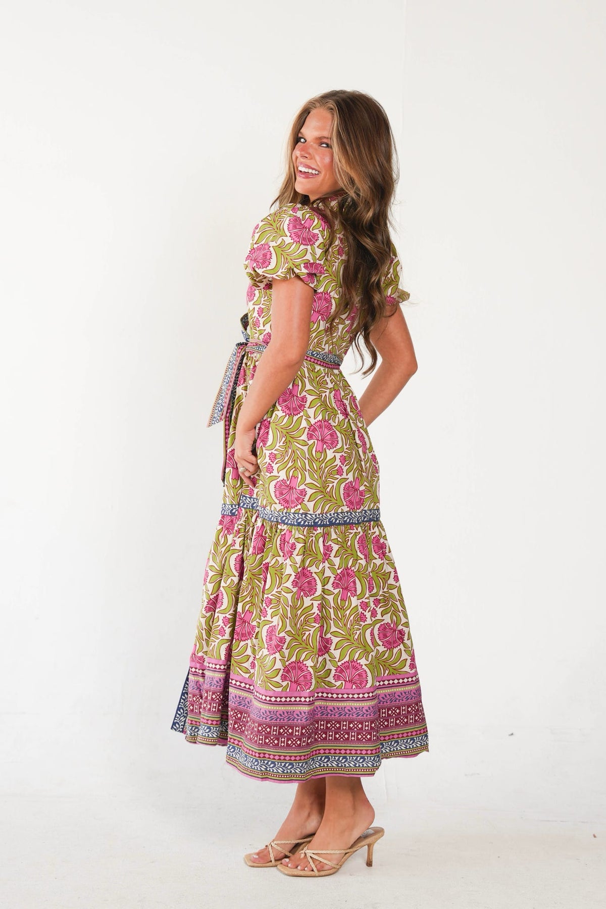 A woman modeling a short-sleeved maxi dress with a vibrant pink and green floral block print, shown from a three-quarters back view against a plain white studio background.