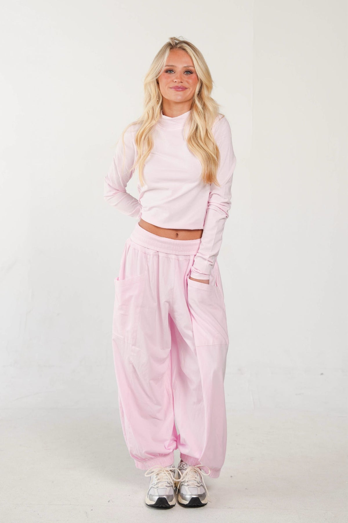 A woman modeling a light pink two-piece lounge set consisting of a long-sleeve cropped top and matching baggy sweatpants, paired with white sneakers, against a plain white studio background.