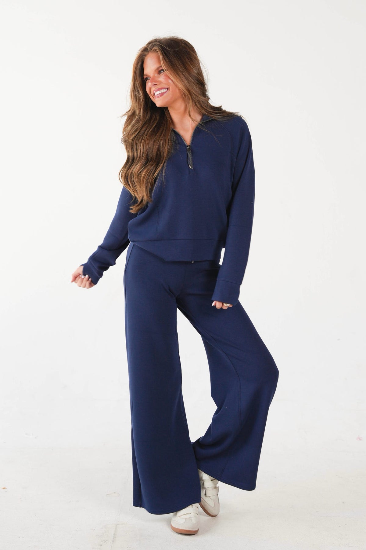 A woman modeling a matching navy blue two-piece lounge set consisting of a long-sleeve quarter-zip top and wide-leg pants, paired with white sneakers against a plain white studio background.
