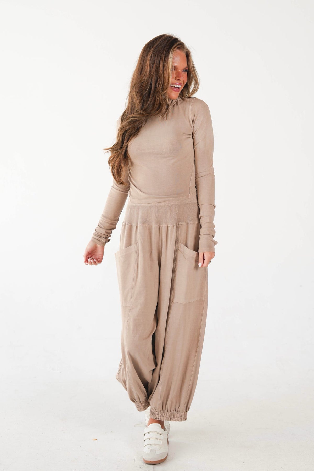 A woman modeling a beige long-sleeve top and matching loose harem-style pants with large front patch pockets, paired with white sneakers, against a plain white studio background.