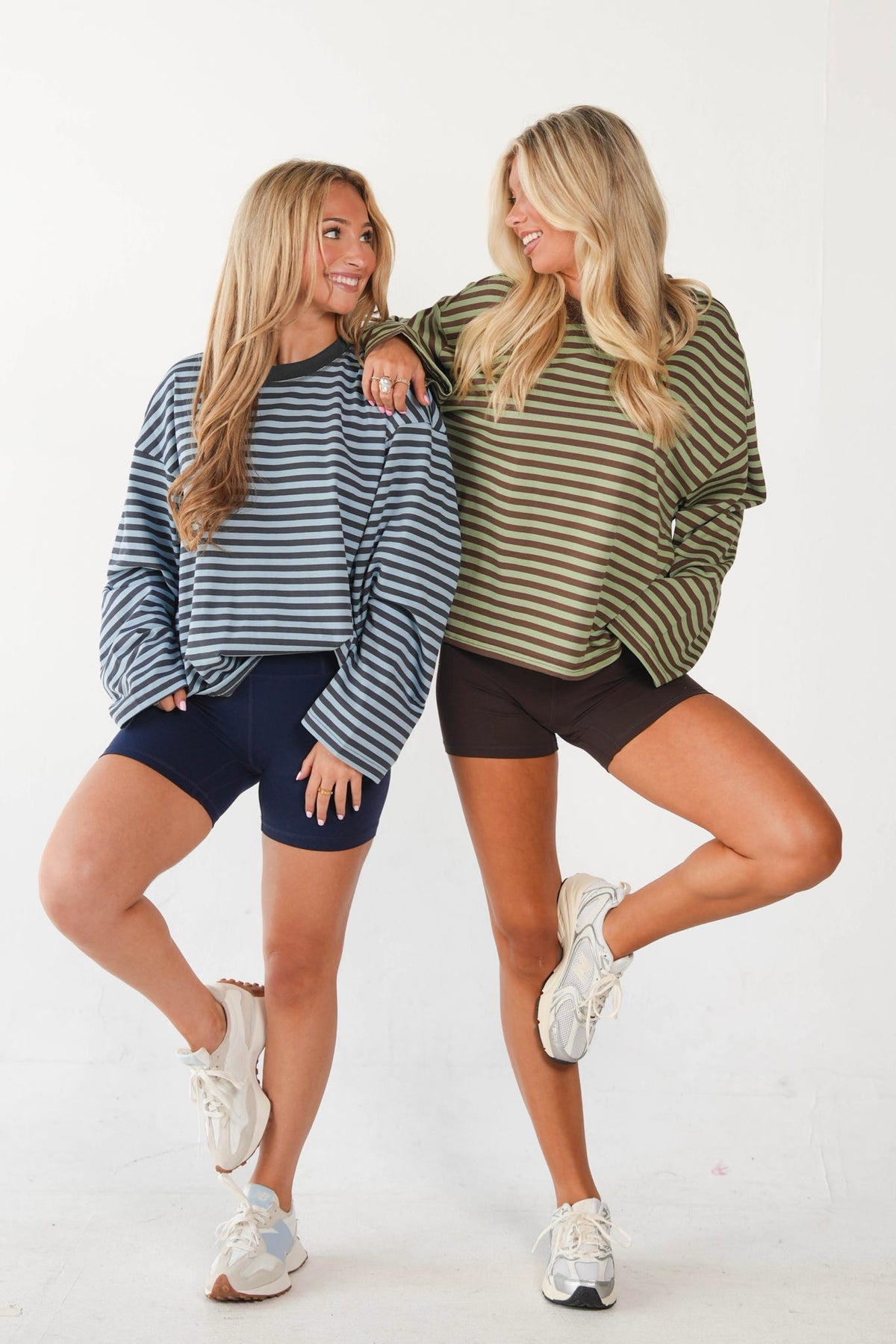 Two women posing together in matching striped long-sleeve crewneck sweatshirts and dark shorts against a plain white studio background.