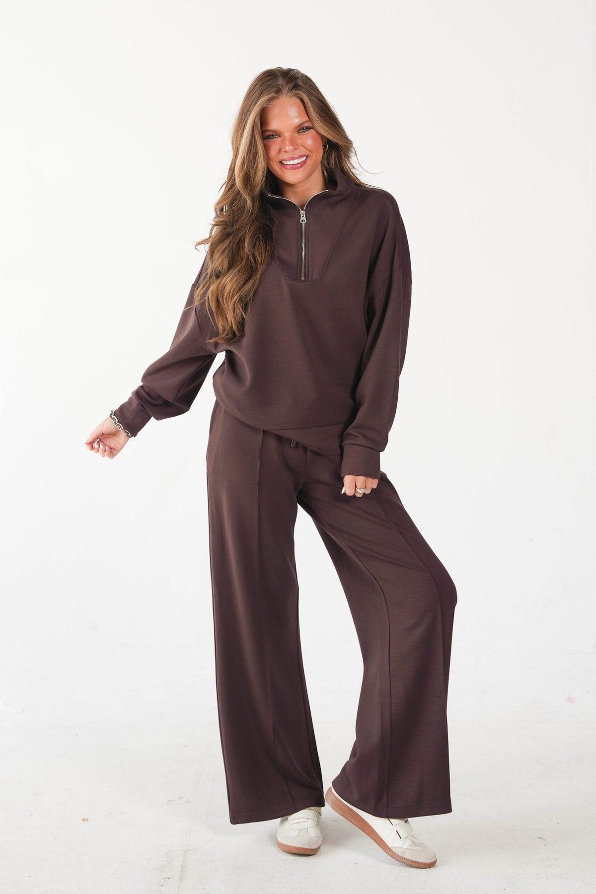 A woman modeling a matching two-piece lounge set in a dark brown color, featuring a long-sleeve quarter-zip pullover and wide-leg pants, paired with white sneakers against a plain white studio background.