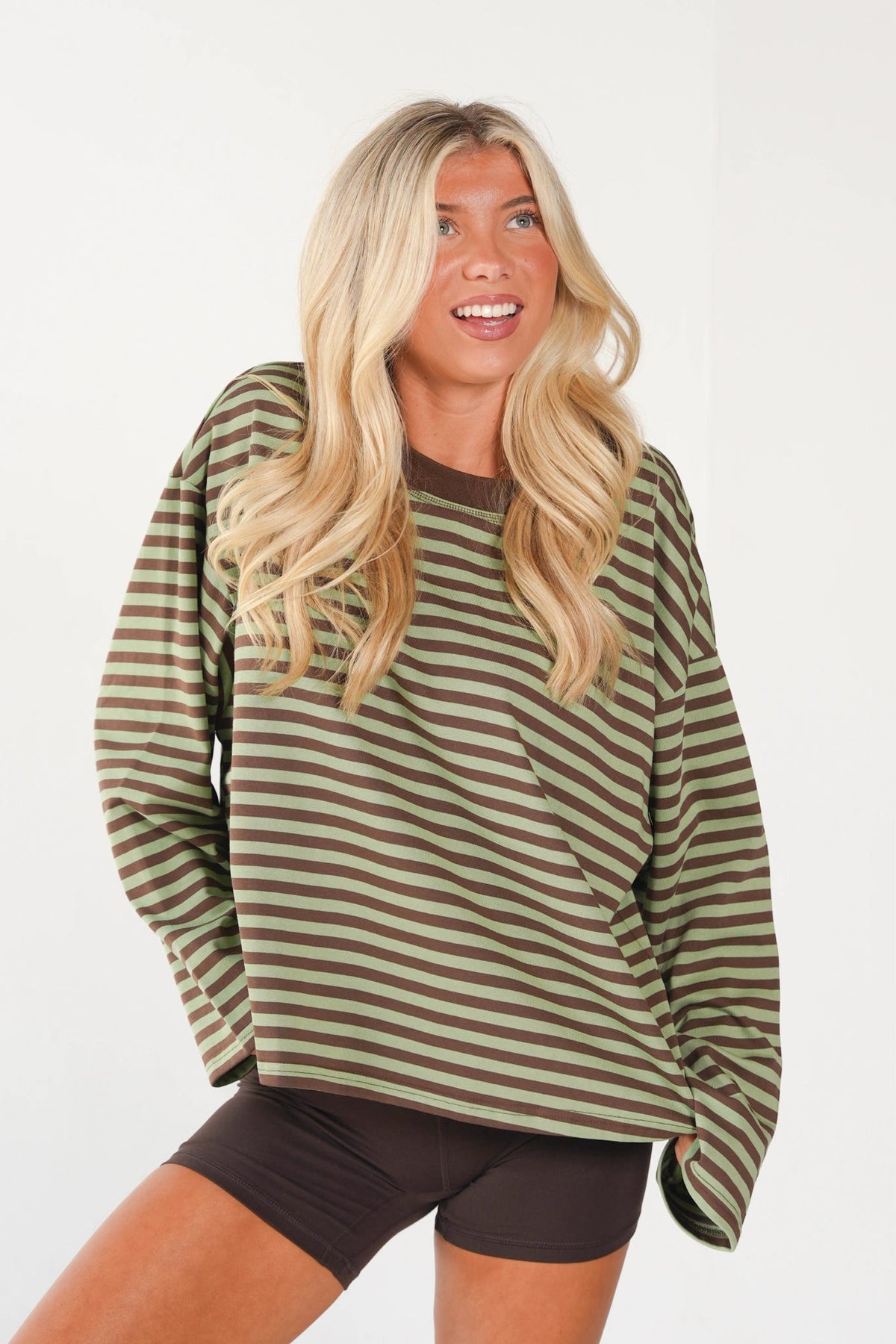 A woman with long blonde hair wearing an oversized long-sleeve shirt with horizontal olive green and brown stripes, paired with dark brown bike shorts, against a plain white studio background.