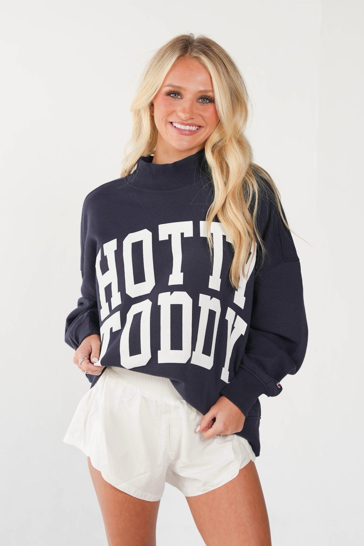 A smiling blonde woman wearing a navy blue mock-neck sweatshirt with 'HOT TOGETHER' printed in bold white letters across the front, paired with white shorts, against a plain white studio background.