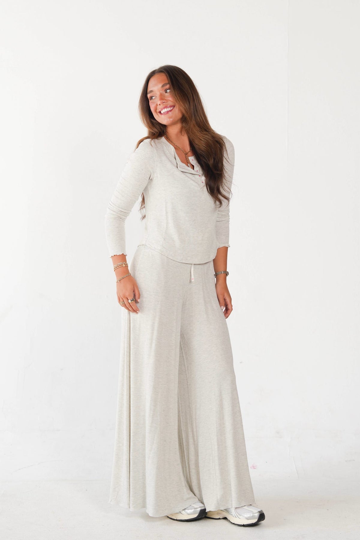 A woman modeling a light beige knit loungewear set consisting of a cowl neck long-sleeve top and matching wide-leg pants, standing against a plain white studio background.