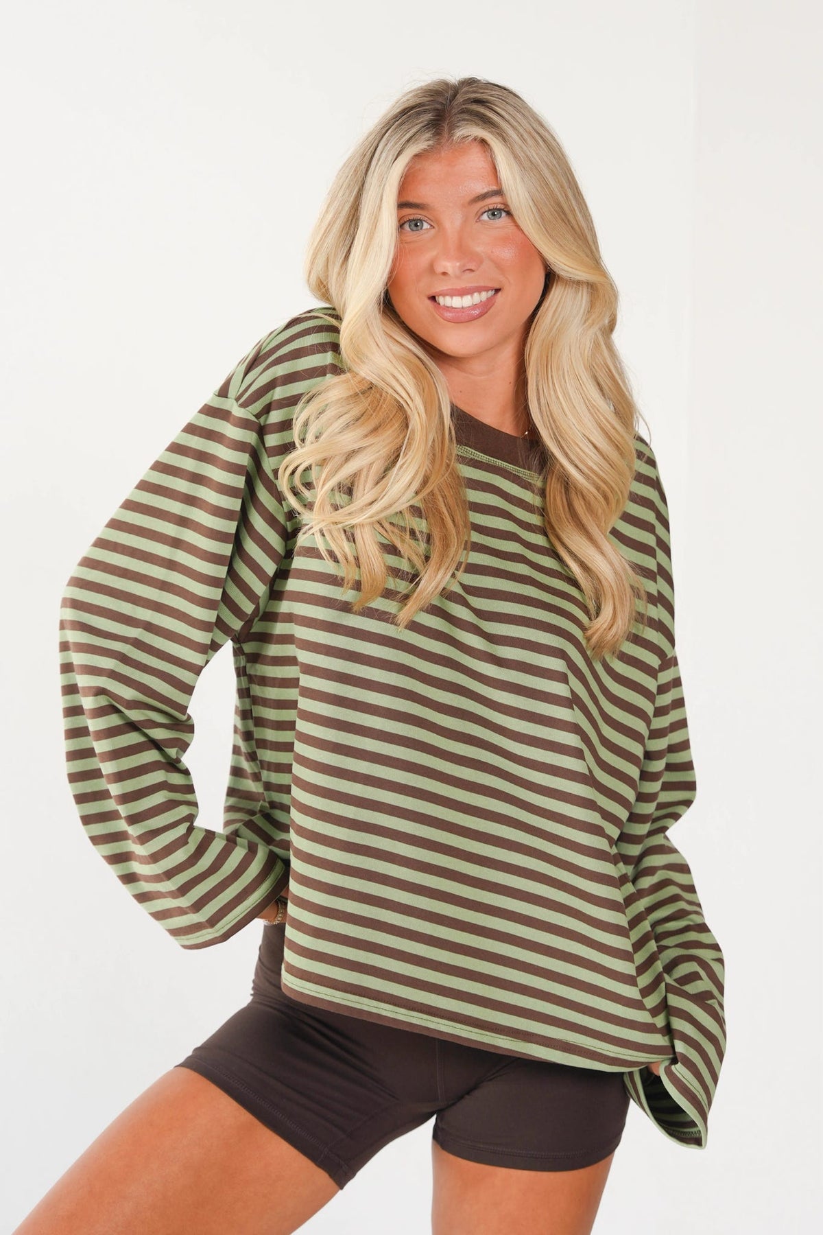 A woman with long blonde hair wearing a long-sleeve crewneck shirt with horizontal olive green and brown stripes, paired with dark brown shorts, posing against a plain white background.
