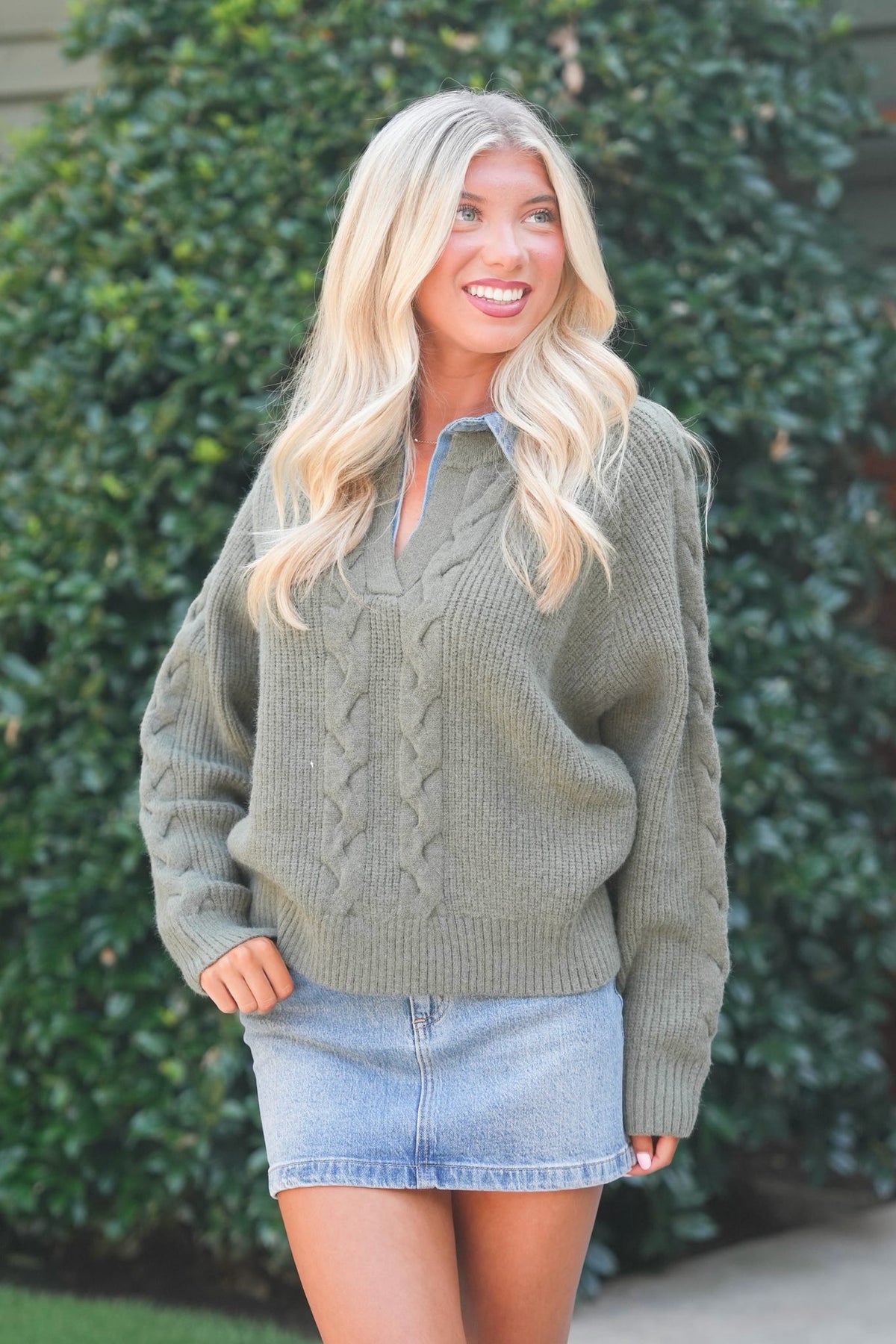 A smiling blonde woman wearing an oversized olive green cable-knit sweater and a light blue denim mini skirt, standing outdoors in front of a green hedge.