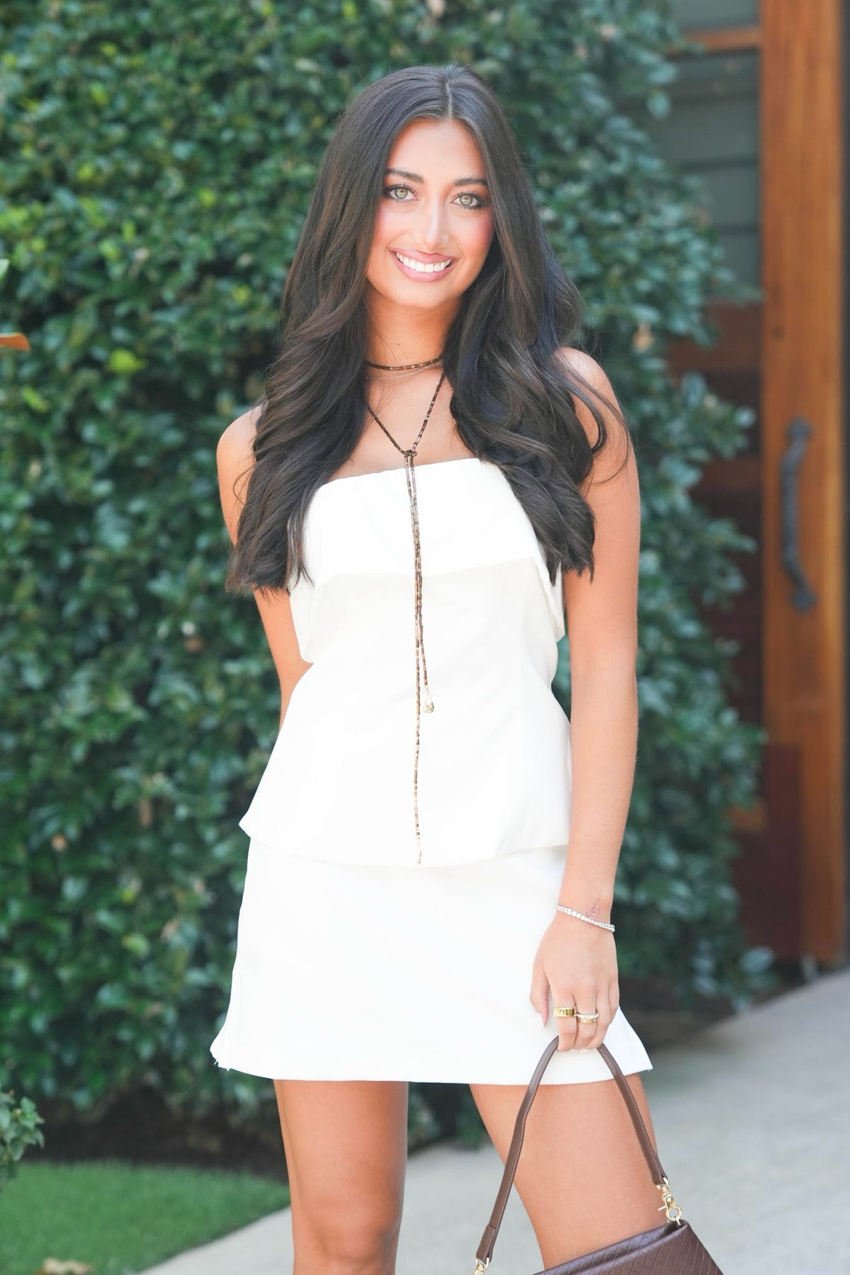A woman with long dark hair stands outdoors in front of a green hedge, wearing a white strapless peplum mini dress with a gold-toned front chain detail. She is holding a brown handbag.