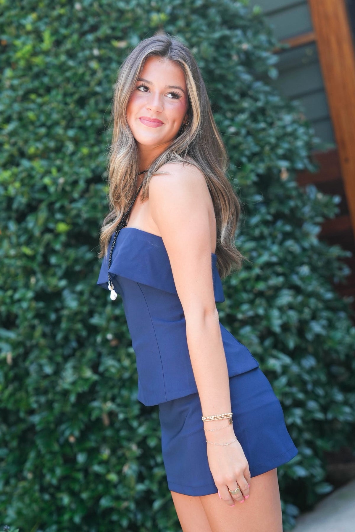 A woman modeling a navy blue off-the-shoulder ruffle mini dress or romper set, standing outdoors in front of a green hedge.