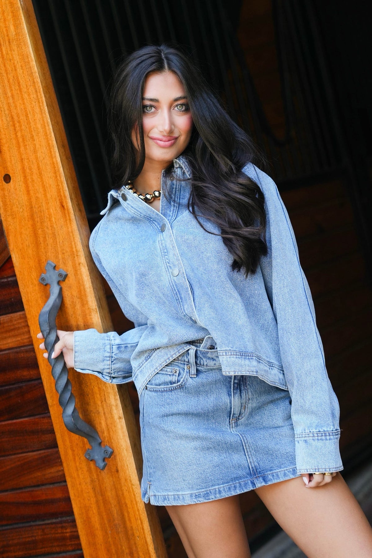 A woman wearing a light blue denim long-sleeve button-up shirt and matching denim mini skirt, leaning against a wooden door frame.