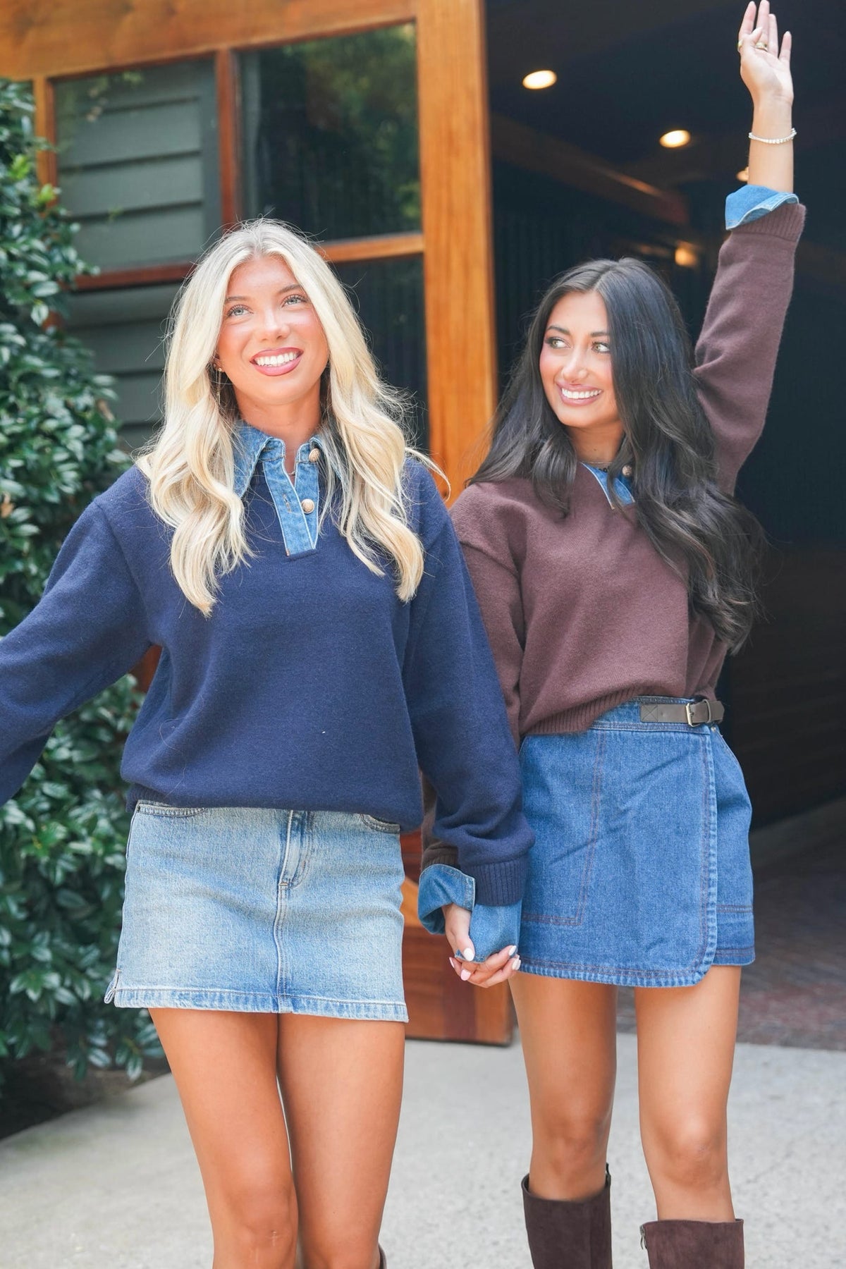 Two women standing outdoors in front of a building entrance, wearing matching navy and mauve sweatshirts with blue denim mini skirts and holding hands.