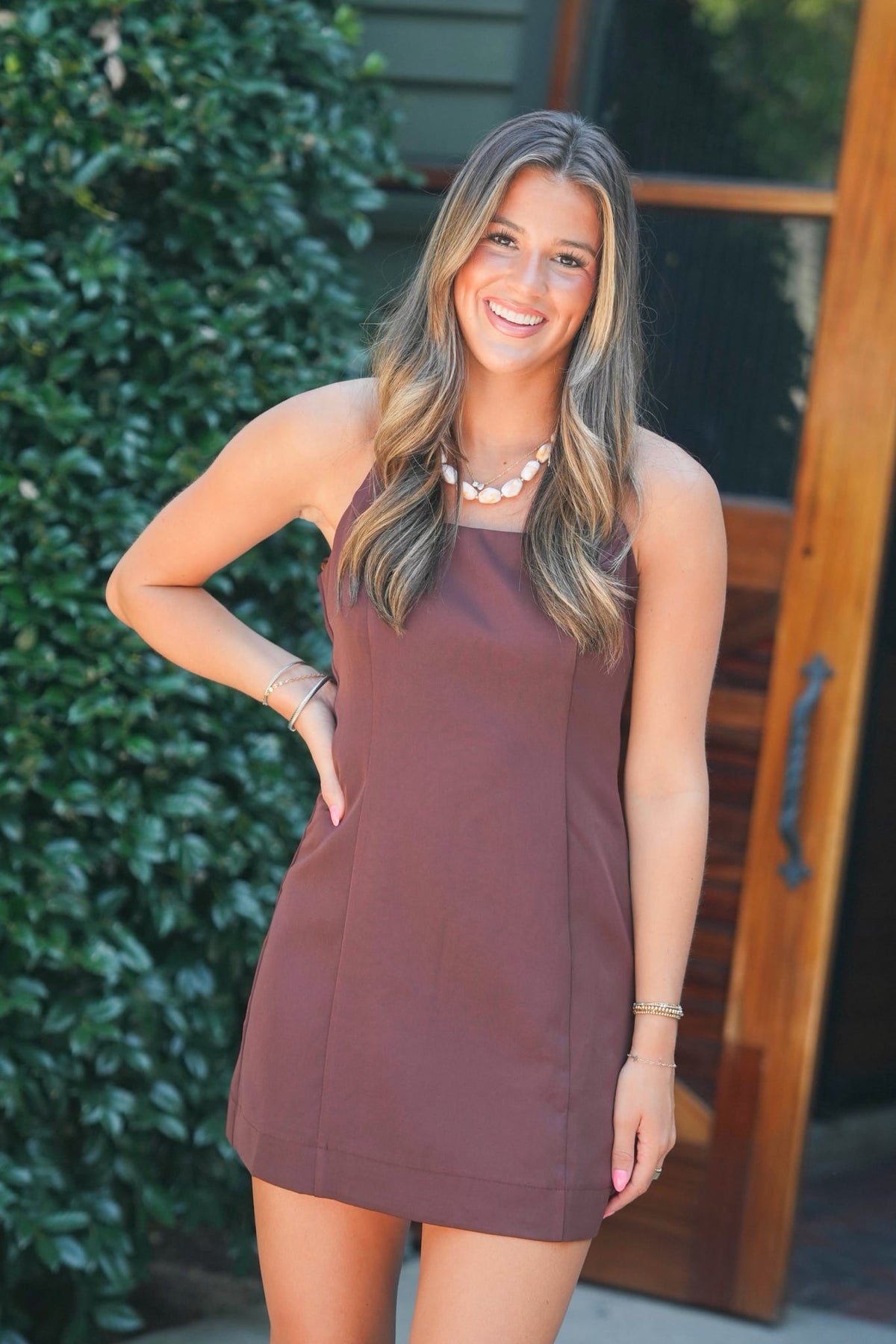 A woman modeling a sleeveless, mini-length dusty rose dress with a square neckline, paired with beige lace-up platform sandals, standing outdoors in front of a wooden door and green foliage.