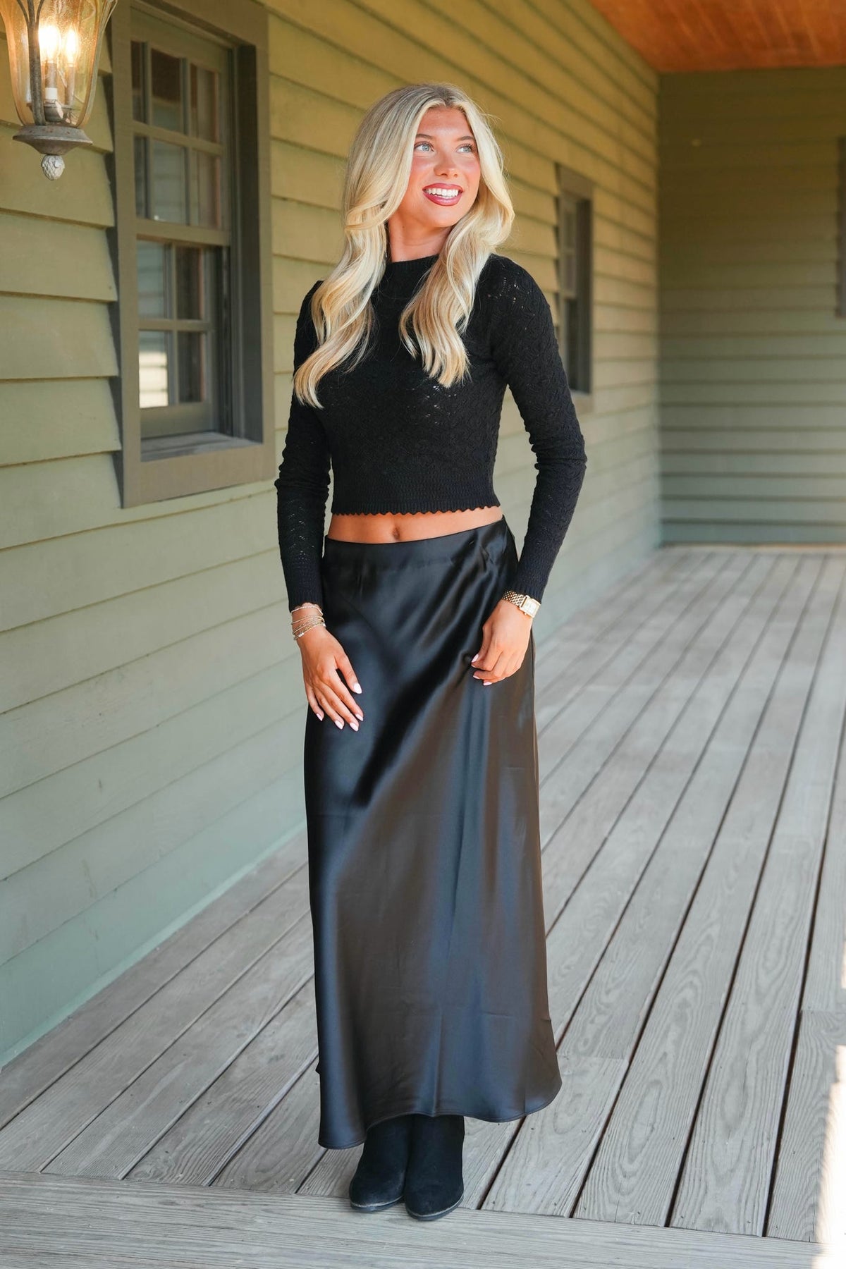 A woman stands on a wooden porch wearing a black long-sleeve knit crop top and a black satin maxi skirt.