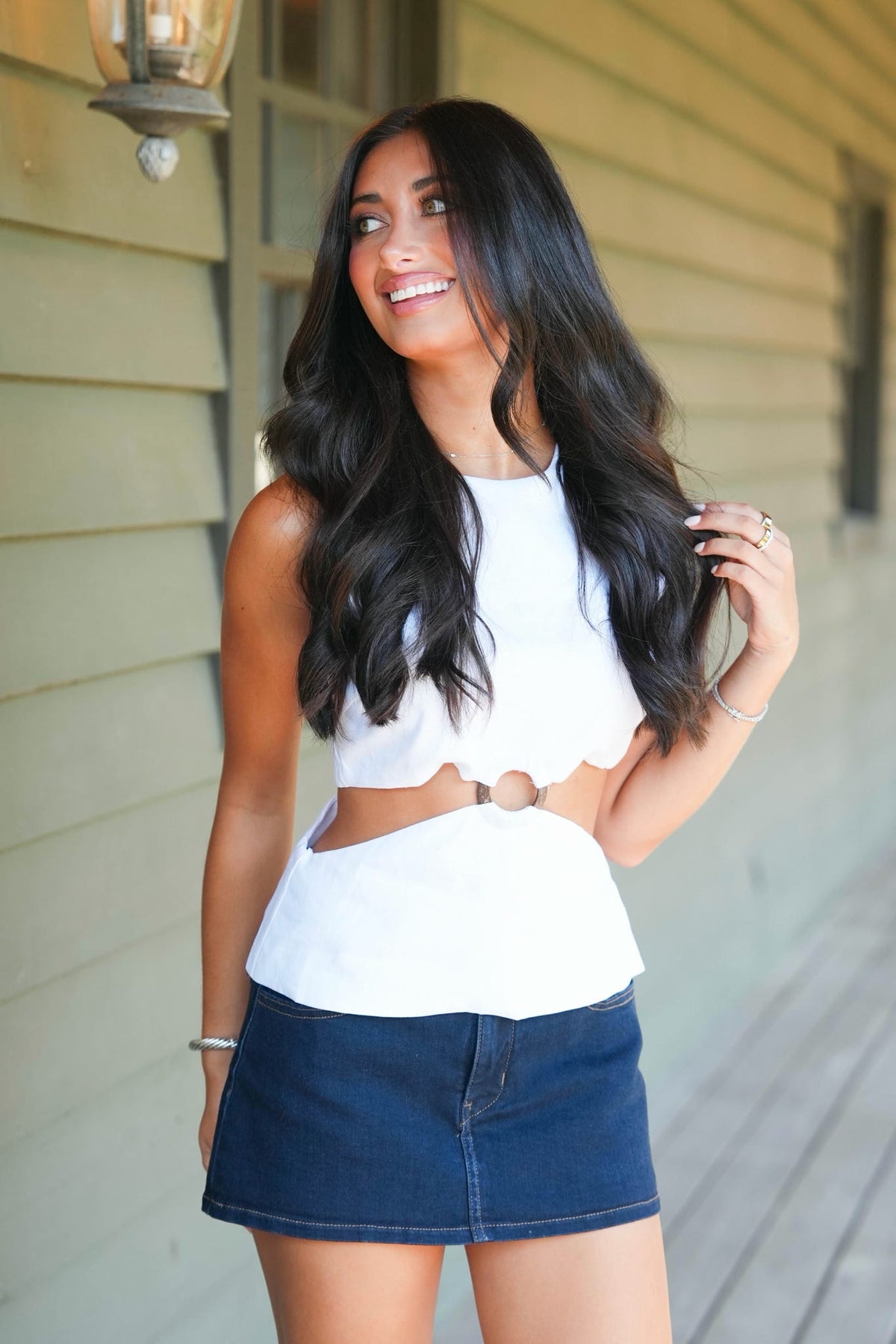 A woman with long, wavy black hair stands on a porch wearing a white sleeveless crop top with a scalloped hem and side cutouts, paired with a dark blue denim mini skirt.