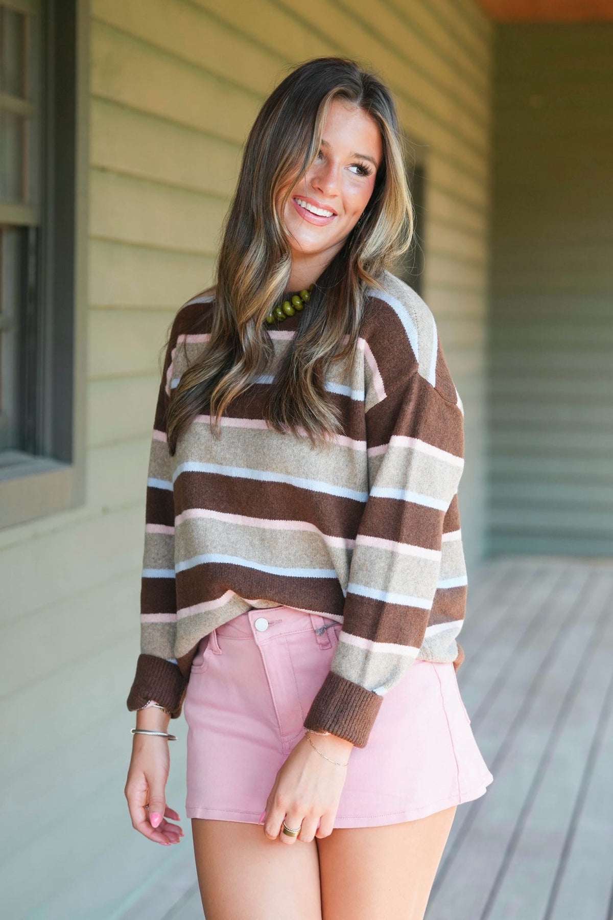 A woman wearing a brown, beige, and white striped knit sweater and a pink mini skirt, standing on a wooden porch.