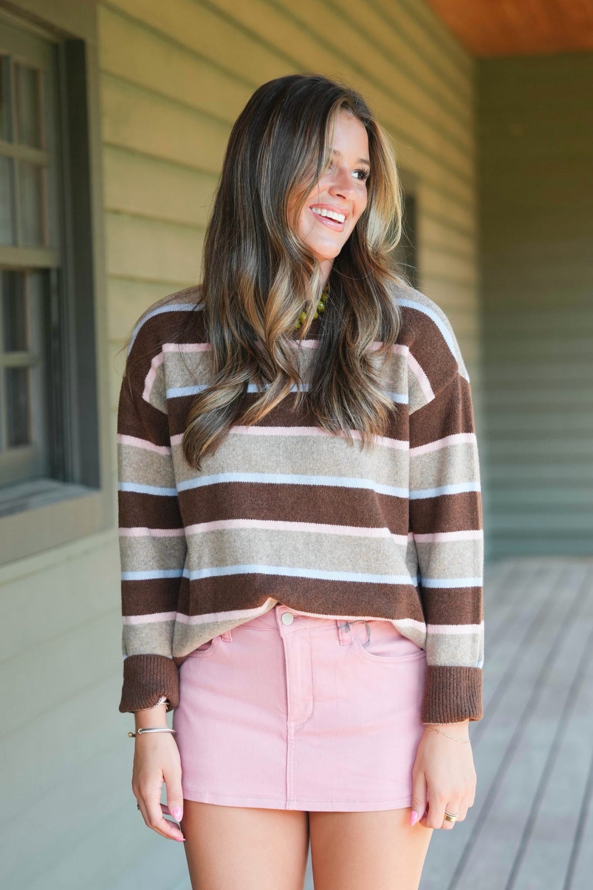 A woman smiling on a porch wearing a brown, beige, and white striped knit sweater and a pink mini skirt.