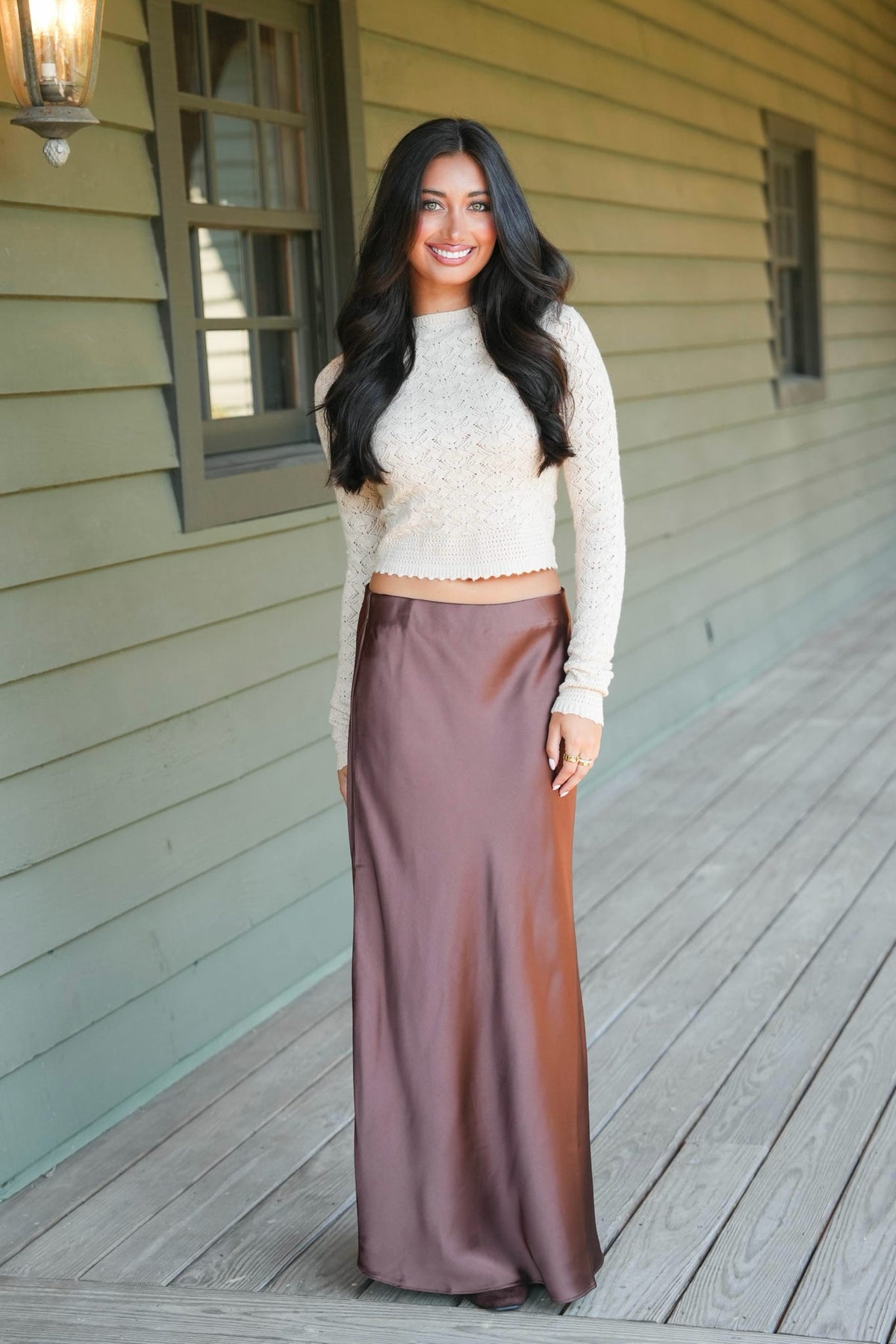 A woman stands on a wooden porch wearing a cream knit long-sleeve crop top and a high-waisted, floor-length brown satin skirt.