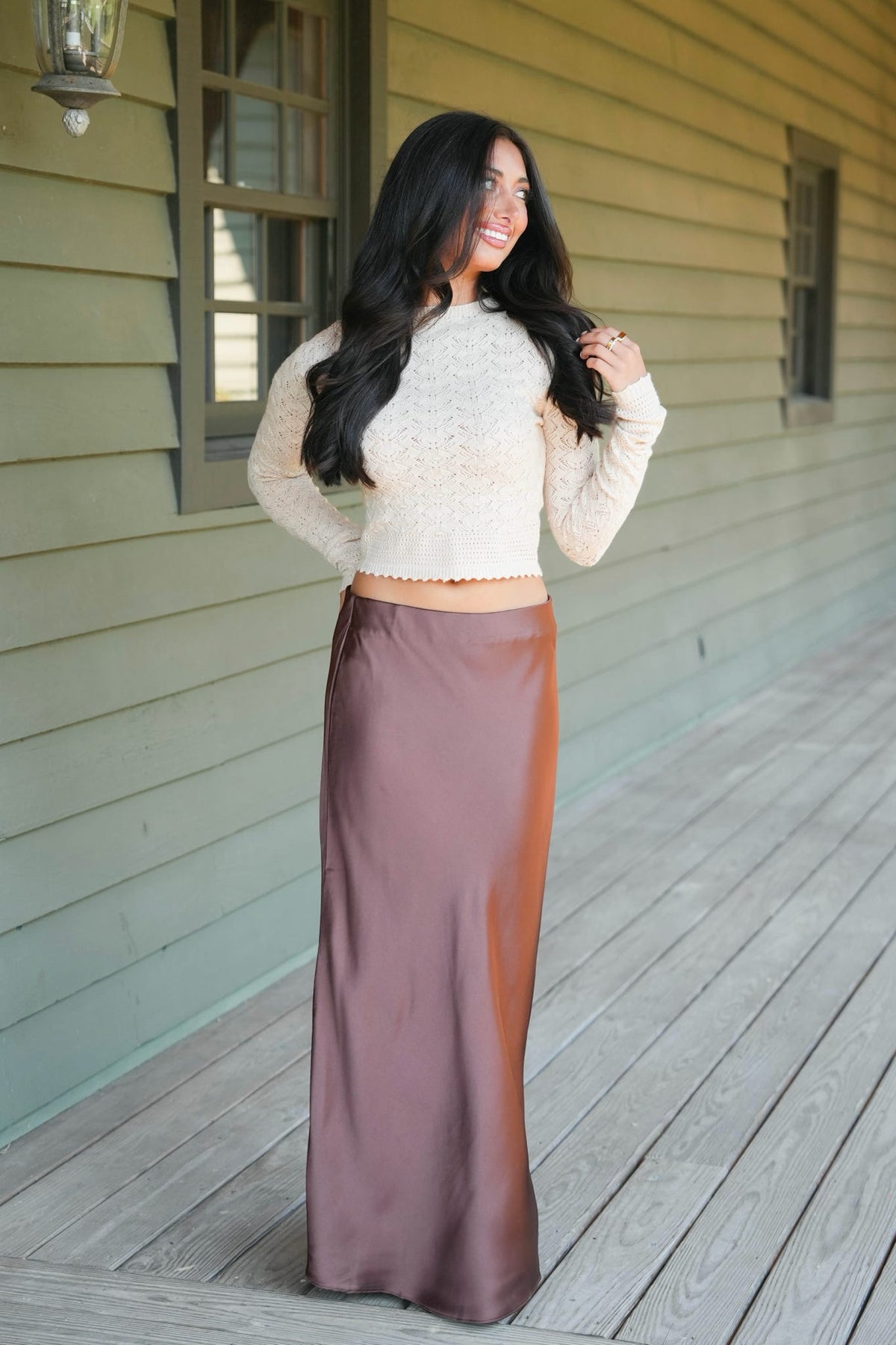 A woman stands on a wooden porch wearing a cream knit cropped sweater and a long mauve-brown satin skirt.