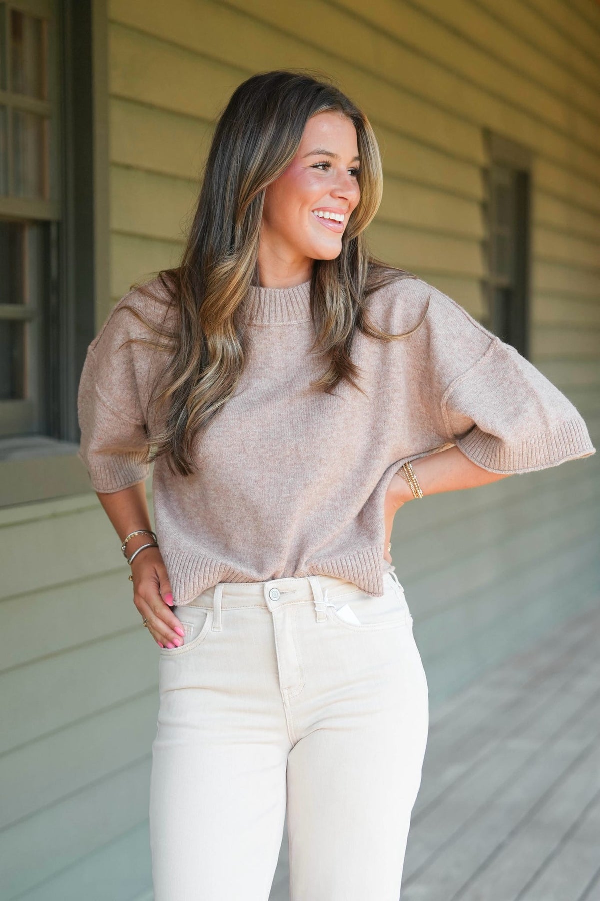 A woman wearing a light beige knit sweater with elbow-length sleeves and white pants, standing on a porch in front of a house with beige siding.
