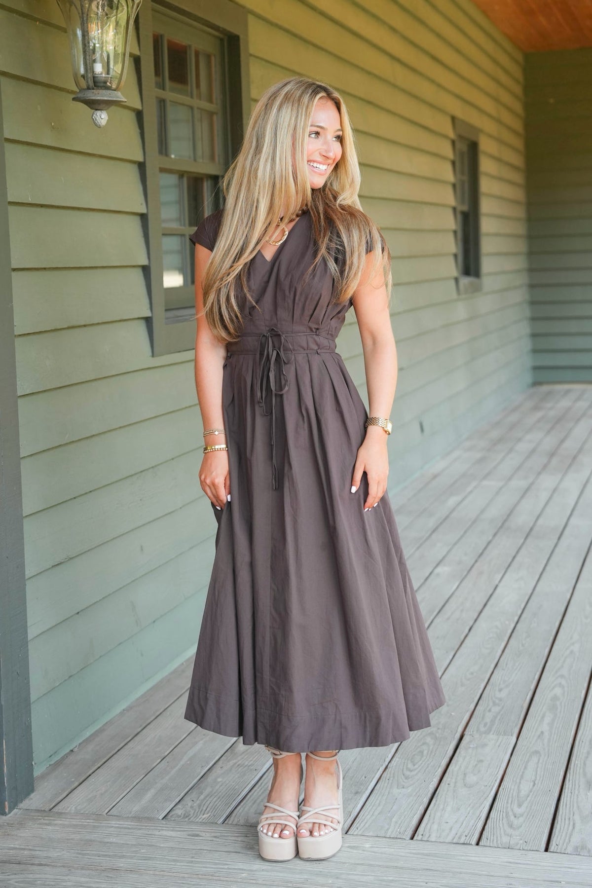A woman stands on a wooden porch wearing a dark brown midi dress with a tie waist and white platform sandals.