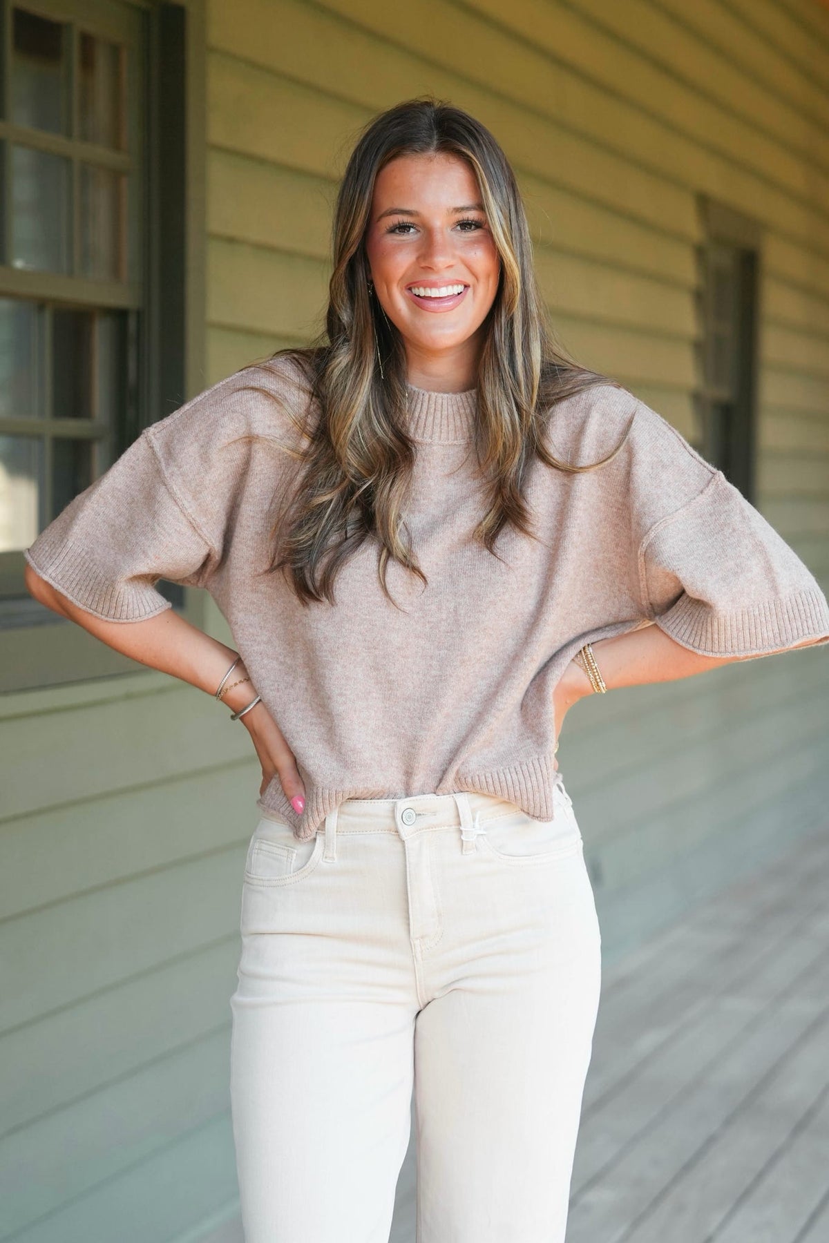 A smiling woman with long brown hair stands on a porch wearing a loose, short-sleeve knit sweater in a light taupe-beige color and off-white jeans.