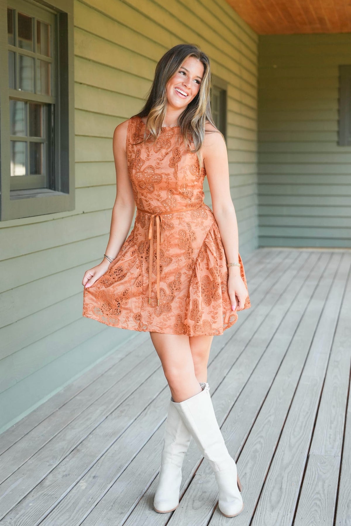A woman stands on a wooden porch wearing a sleeveless rust-orange patterned dress with a tie waist and white knee-high boots.